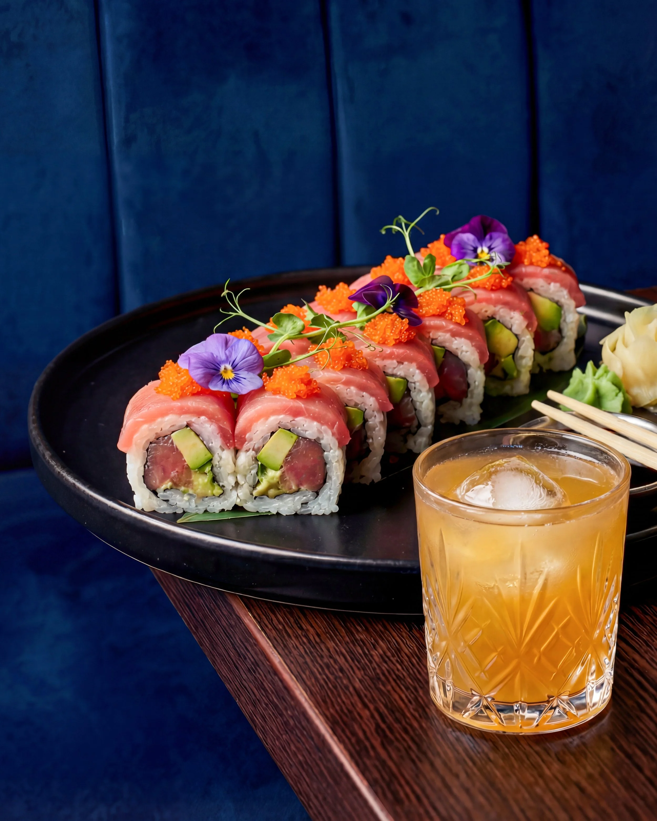A black platter of sushi rolls garnished with edible purple flowers and orange fish roe, with a glass of amber-colored cocktail on a wooden table.