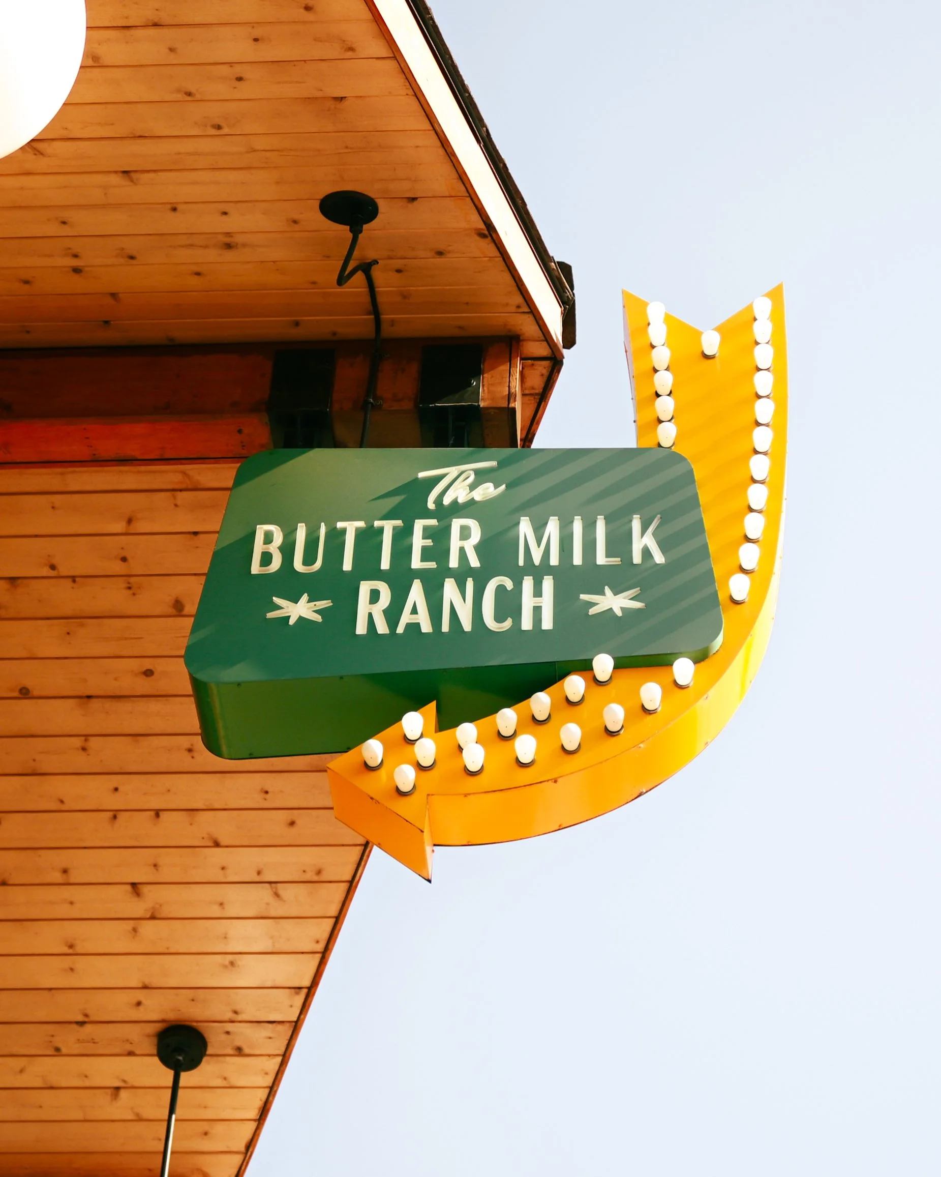 A green sign with white text reading 'The Butter Milk Ranch' hangs from a wooden building with a triangular yellow sign and a border of round bulbs. The sky is visible in the background.