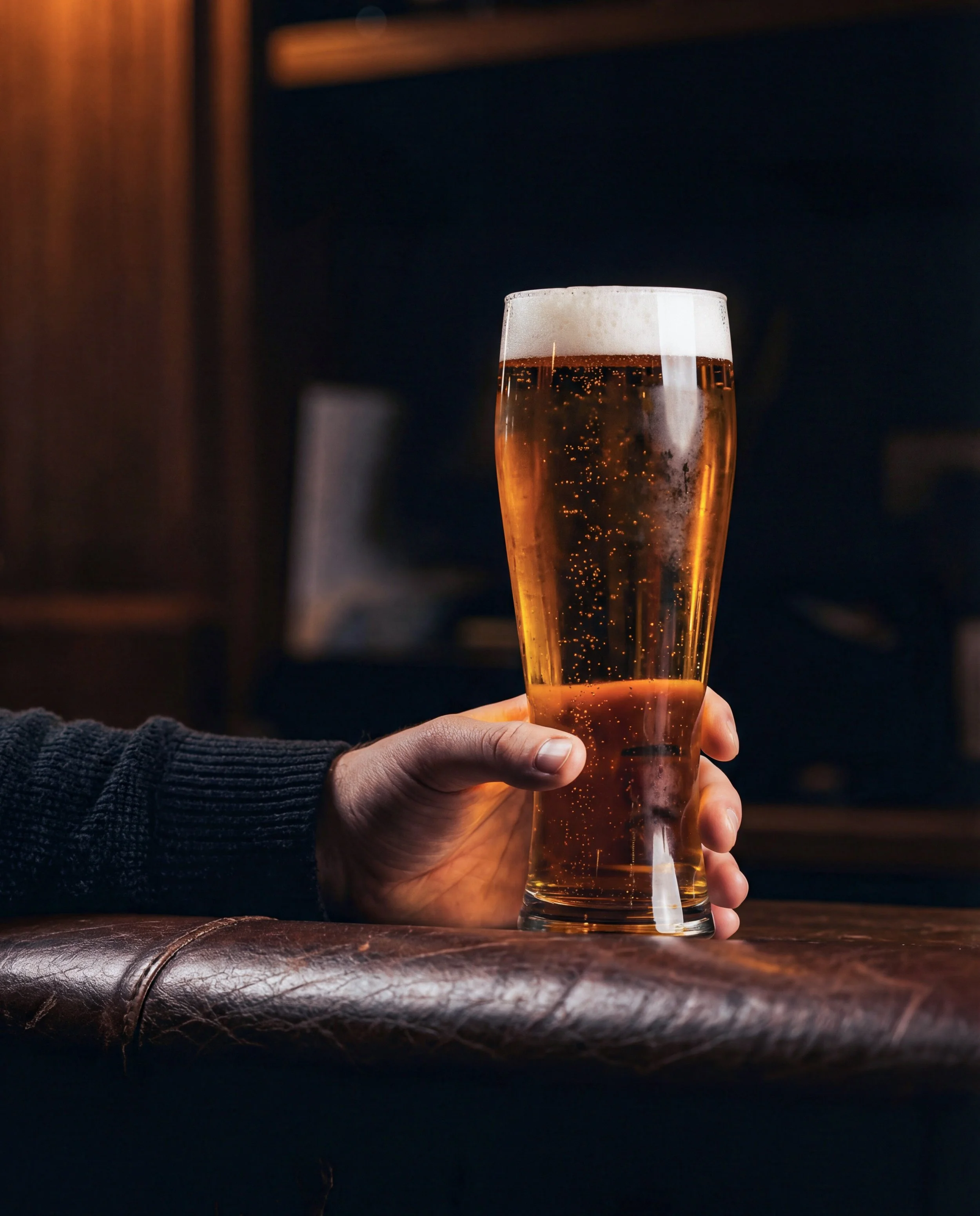 Person holding a pint glass of beer with a foamy head in a dimly lit setting.