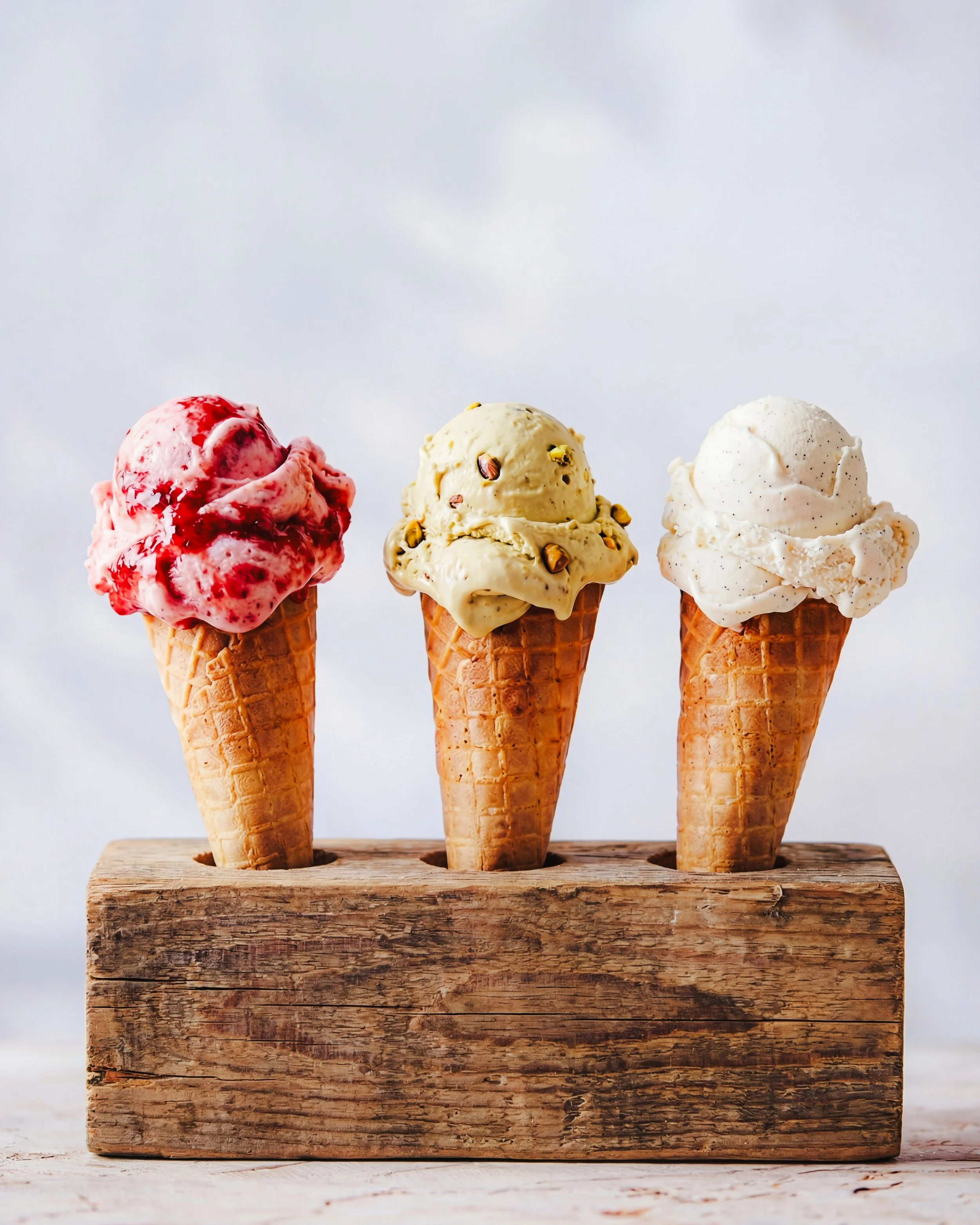 Three scoops of ice cream in waffle cones, placed in a wooden holder. The ice cream flavors are strawberry, pistachio, and vanilla. The background is a soft, cloudy sky.