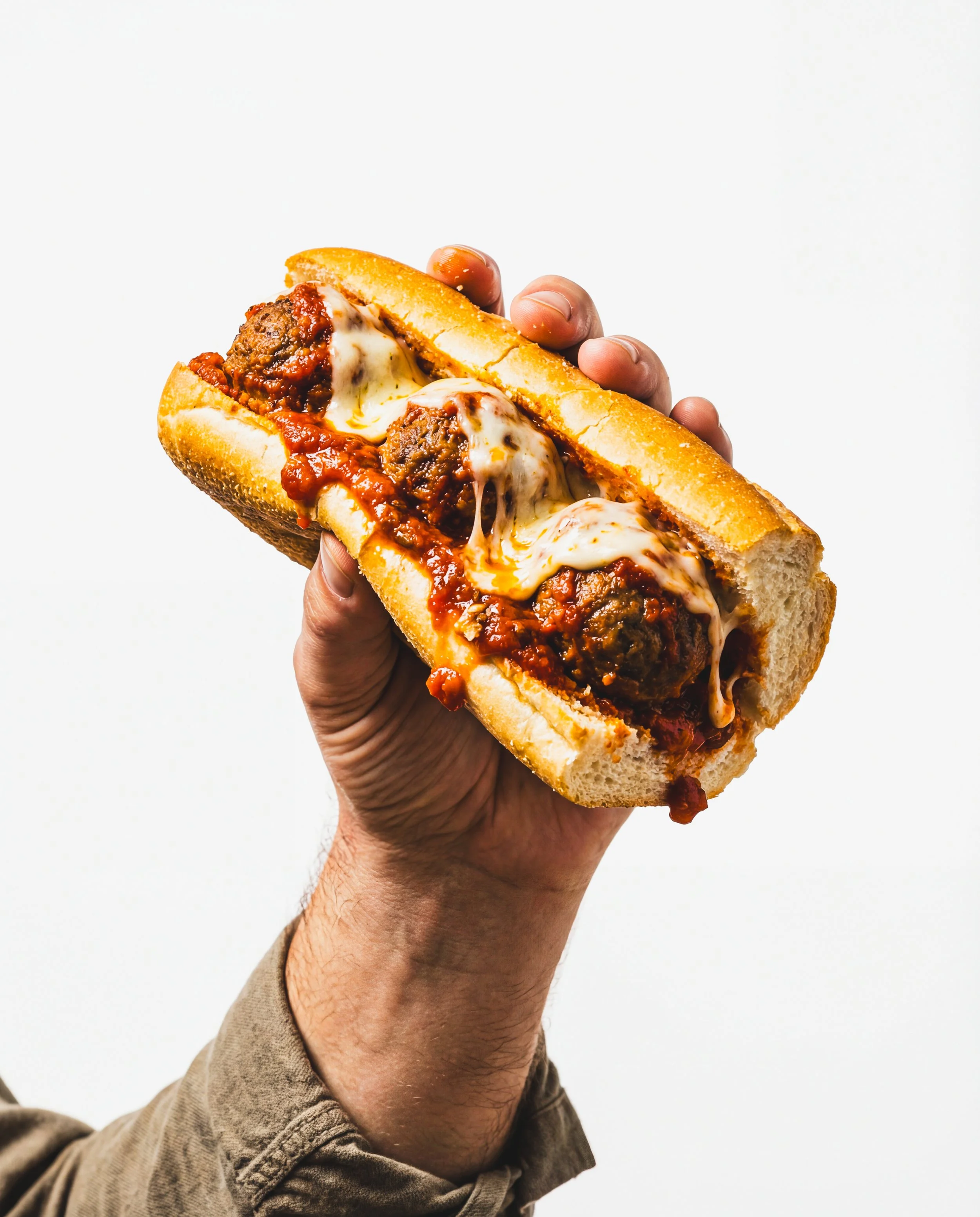 Close-up of a person holding a large meatball sub sandwich with marinara sauce and melted cheese, against a white background.