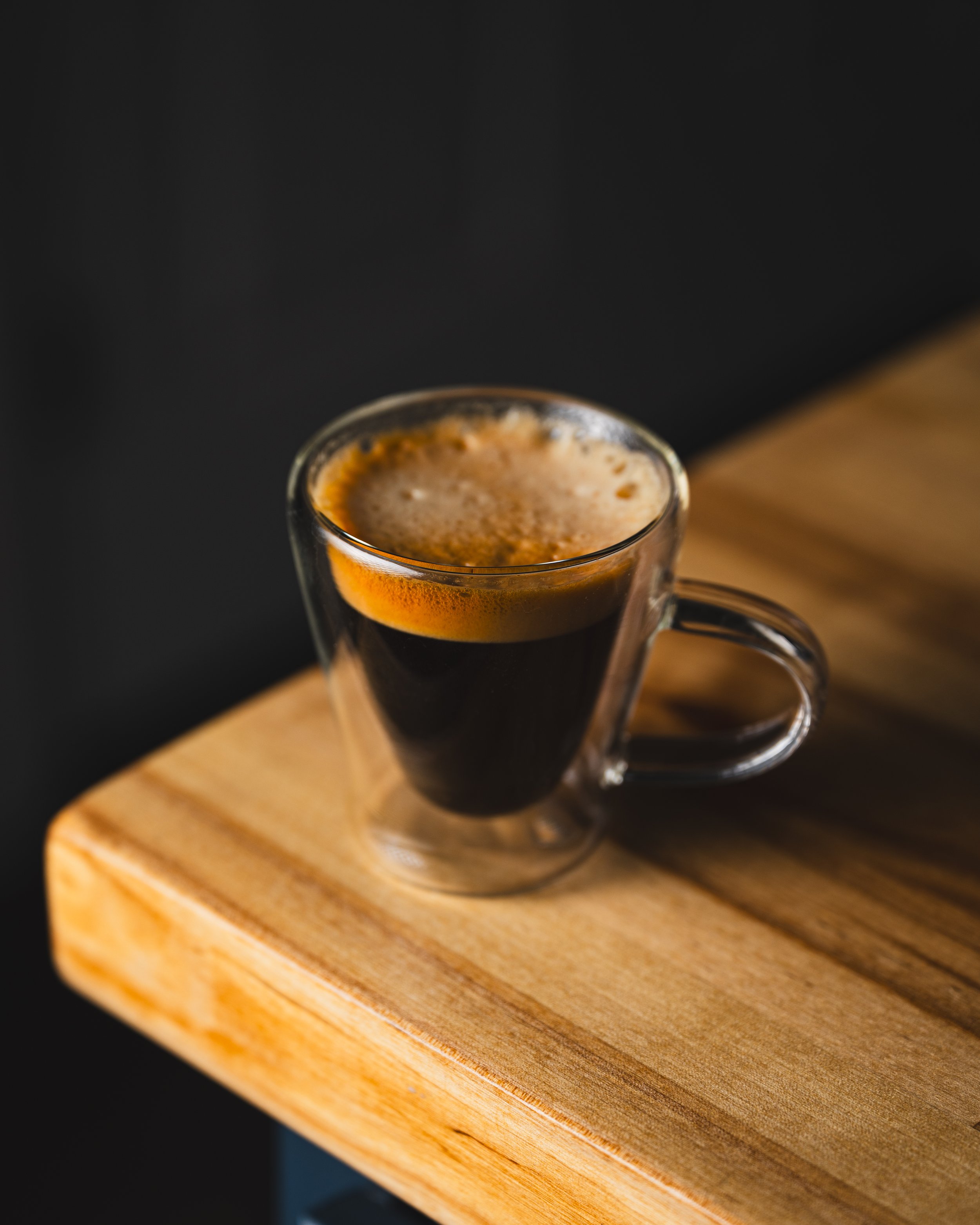 A clear glass coffee cup filled with black coffee, sitting on a wooden table.