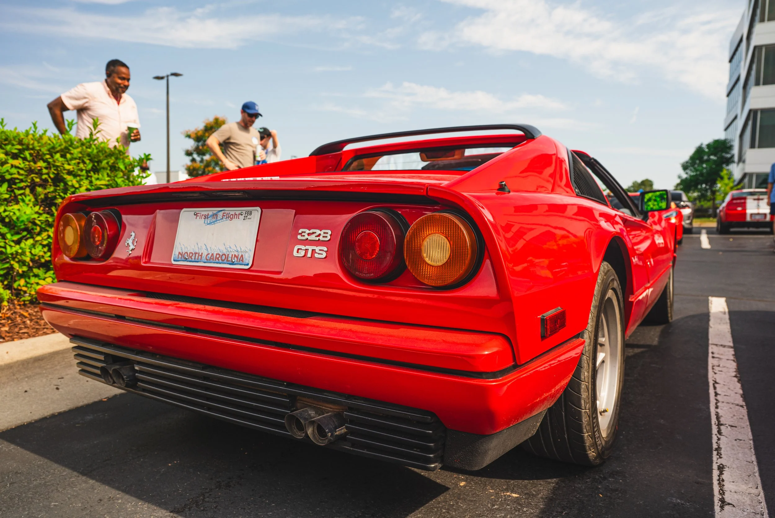 A red Ferrari 328 GTS sports car parked in a parking lot with a North Carolina license plate. People are standing nearby, some holding drinks, in an outdoor setting with buildings and trees in the background.