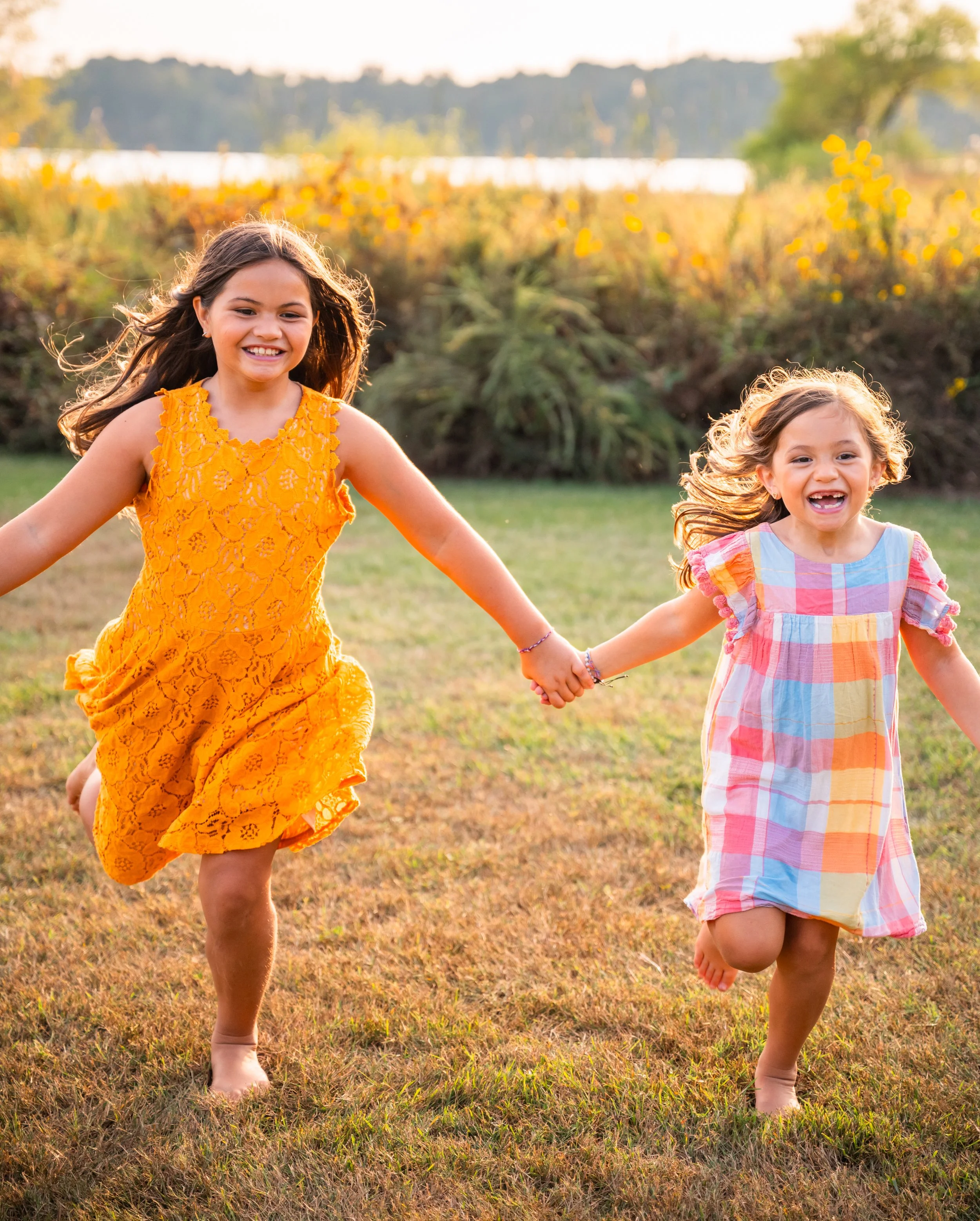 Two young girls holding hands and running outdoors on a grassy field during sunset, wearing colorful dresses, with trees and a body of water in the background.