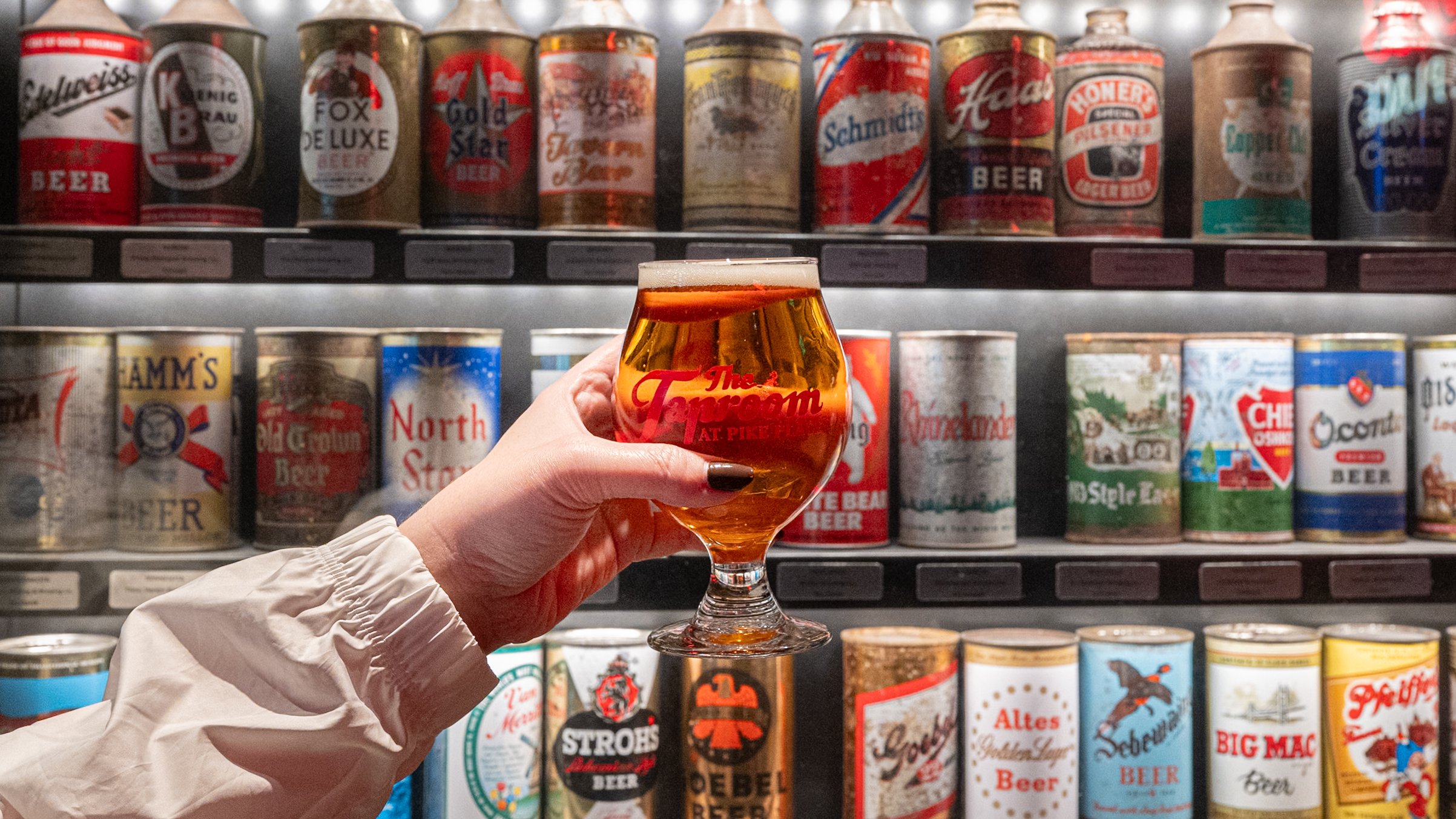Person holding a tulip-shaped beer glass filled with golden beer in front of a display of vintage beer cans on store shelves.