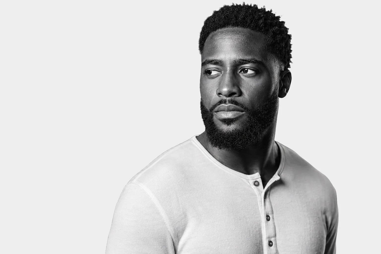 Black and white portrait of a man with a beard and curly hair, wearing a light-colored, buttoned henley shirt, looking to the side with a neutral expression.