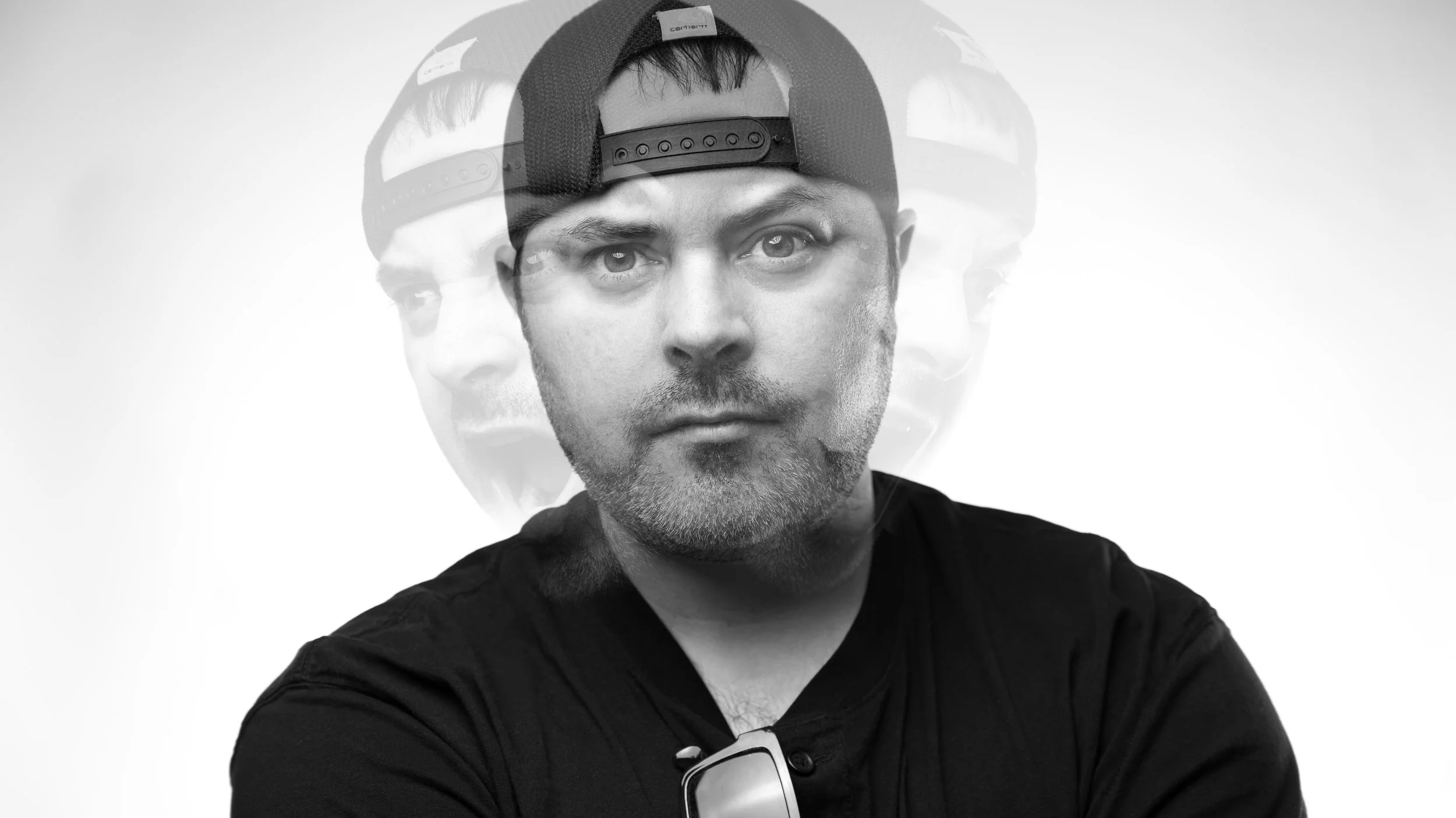 Black and white photo of a man with a beard wearing a backwards cap and black t-shirt, with a double exposure effect showing multiple overlapping images of his face.