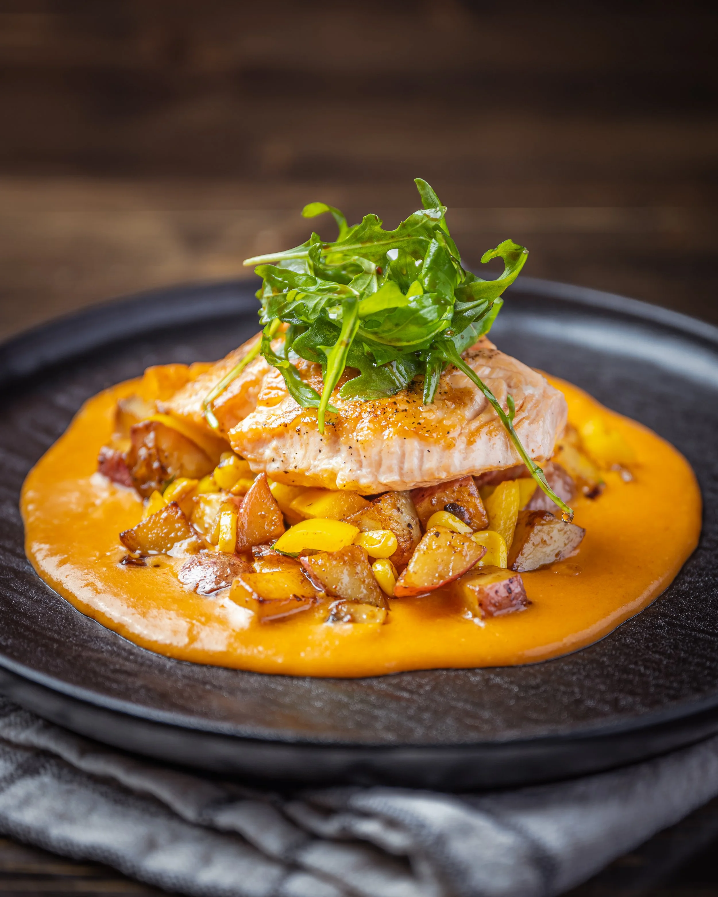 A plated dish of cooked salmon topped with fresh greens, served over a bed of roasted vegetables and a smooth orange sauce on a black plate.