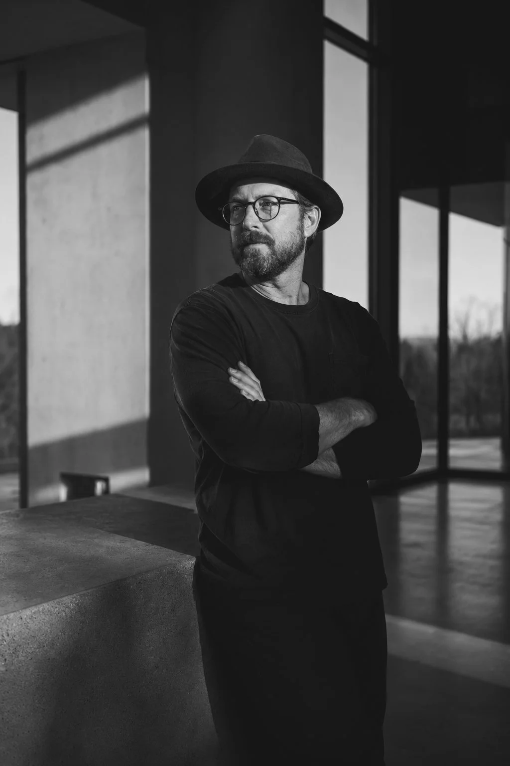 A man wearing glasses, a hat, and a black shirt standing indoors with arms crossed, near large windows with sunlight streaming in.