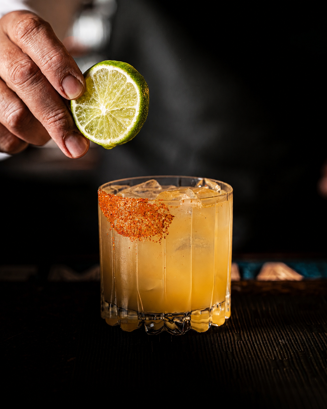 A hand squeezing a lime wedge over a glass of cocktail with ice and a salted rim.