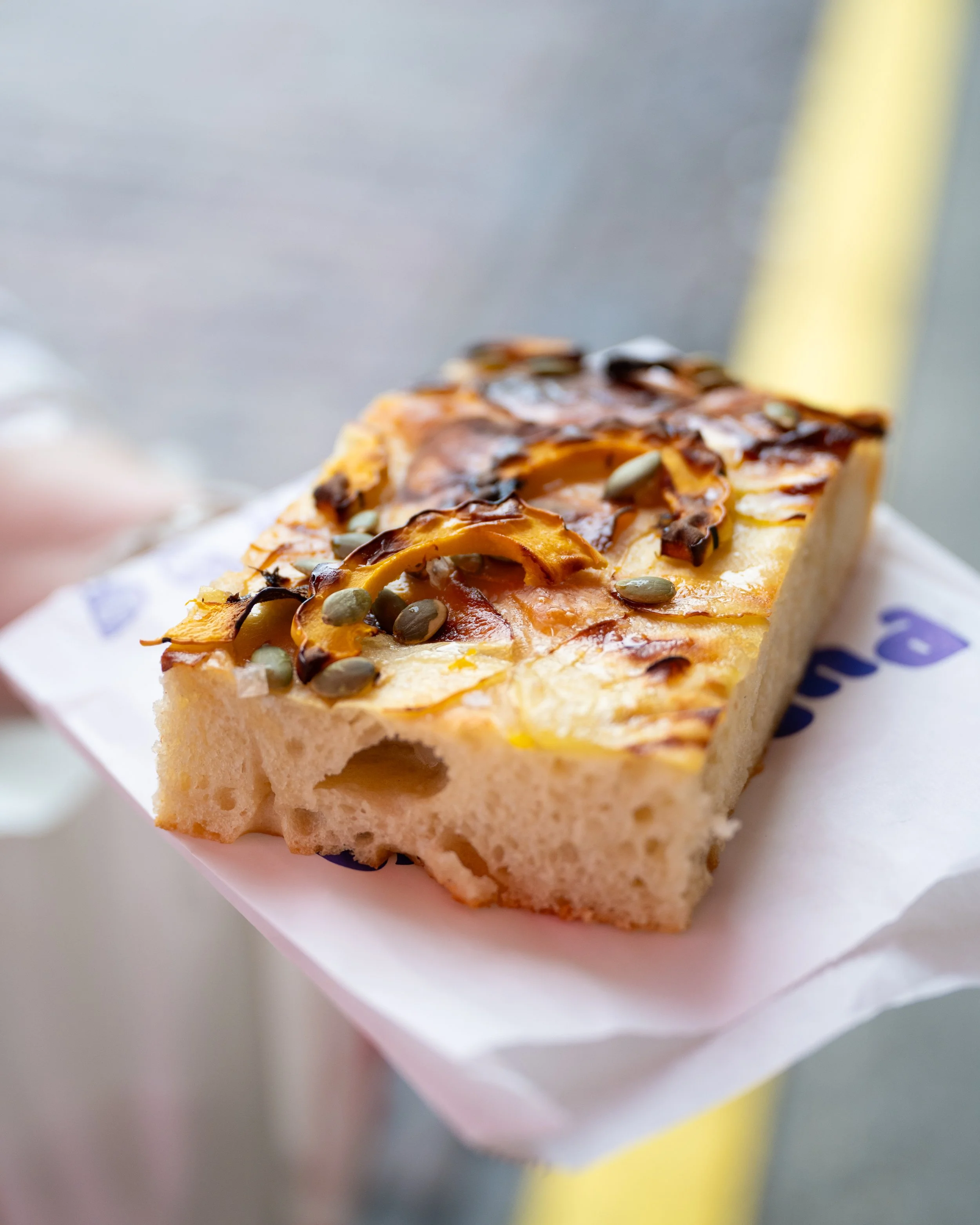 A slice of focaccia bread topped with roasted vegetables and pumpkin seeds.
