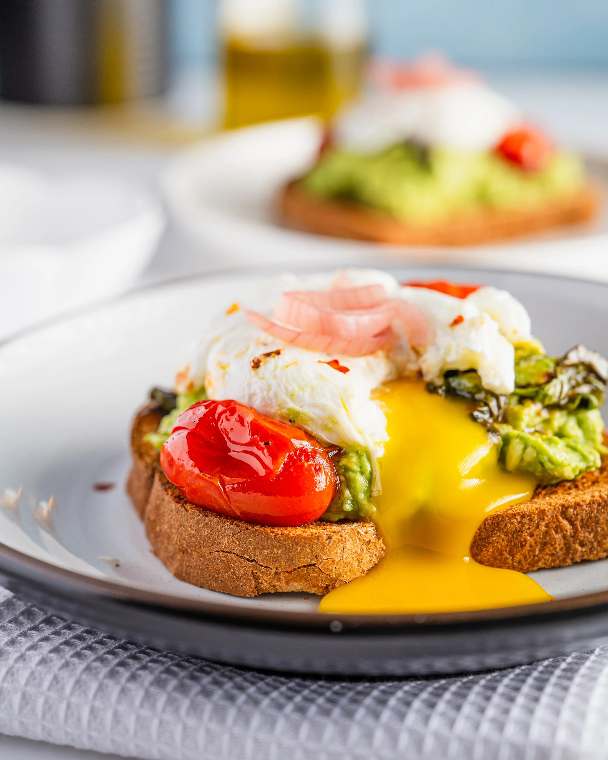 Close-up of toast with avocado, cherry tomatoes, poached egg, and partially broken runny yolk, served on a white plate.