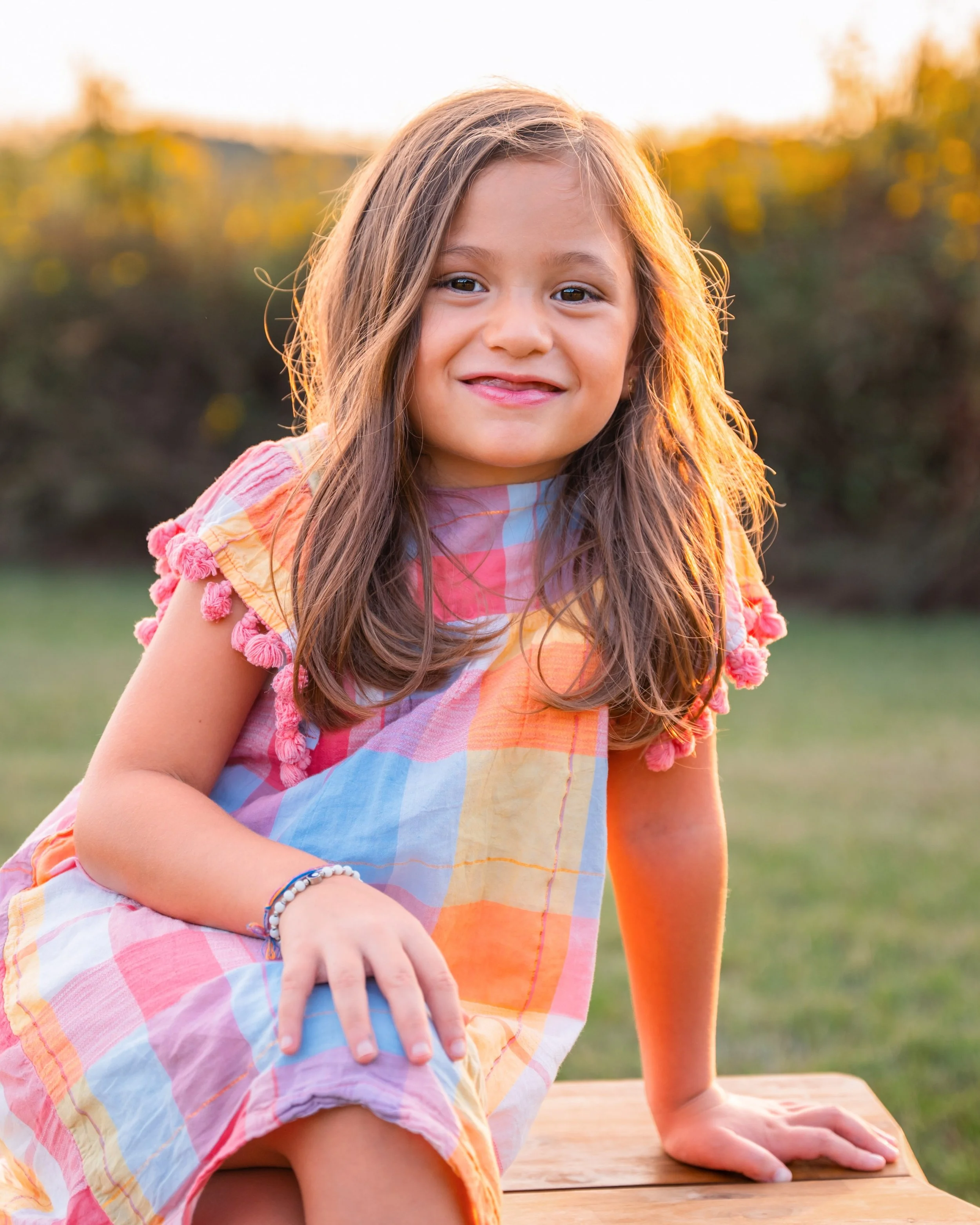 A young girl with long, wavy brown hair smiling, sitting outdoors on a wooden surface during sunset, with yellow-orange tones in the background.