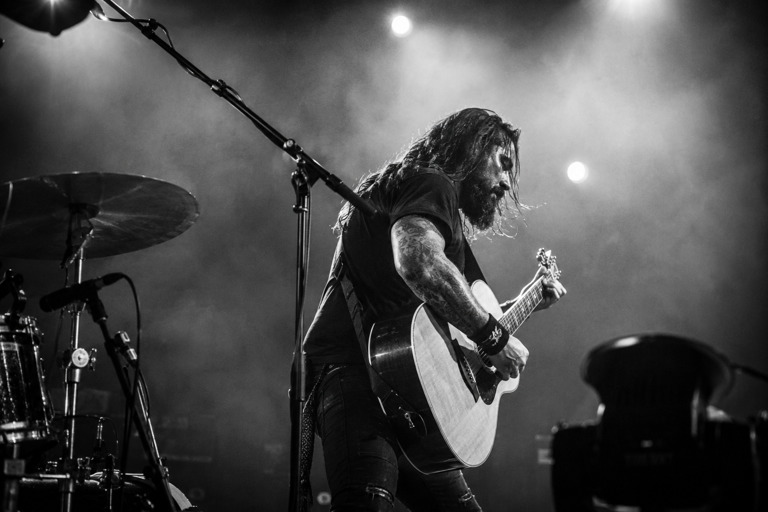 A male musician with long hair and tattoos playing an acoustic guitar on stage, with drums and microphones visible, under stage lighting.