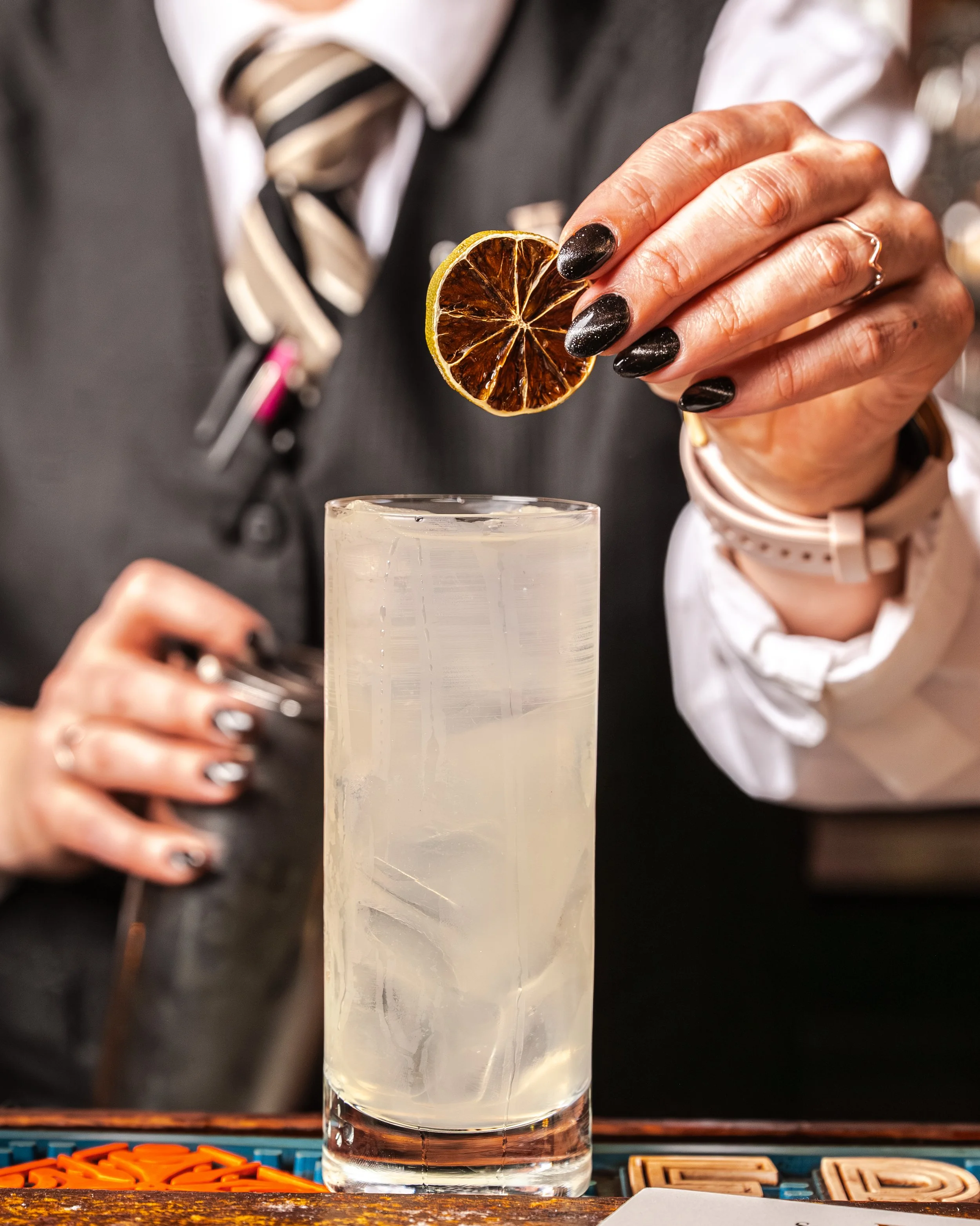 A close-up of a hand holding a dried citrus slice over a glass of clear liquid, with a bartender in a white shirt, gray vest, and striped tie in the background.