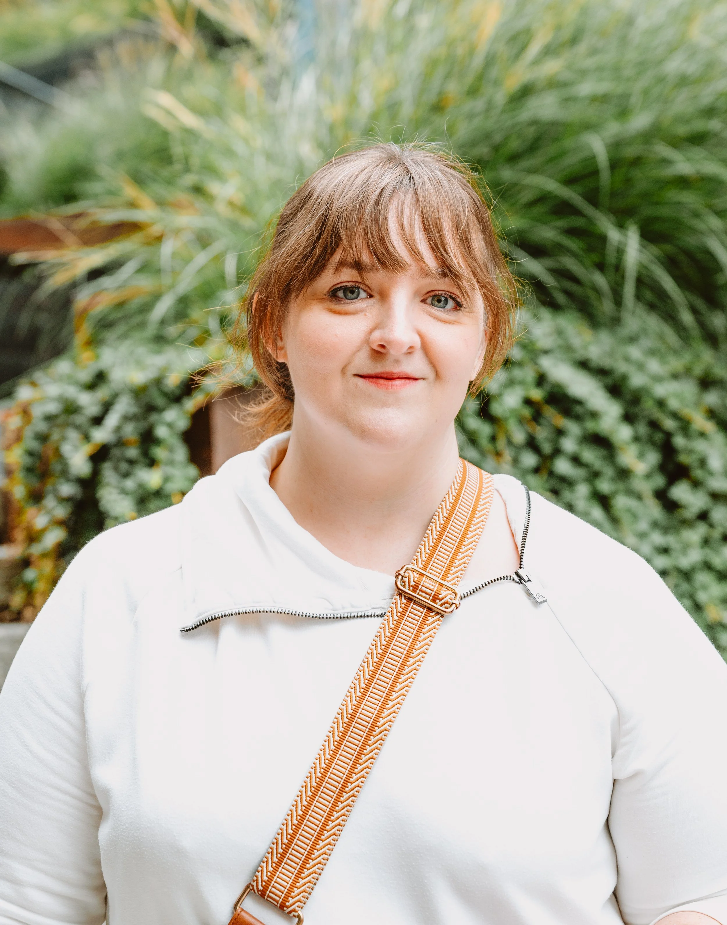 A woman with shoulder-length brown hair and blue eyes standing outdoors with green bushes and plants in the background, wearing a white jacket with a zipper and a beige crossbody strap.