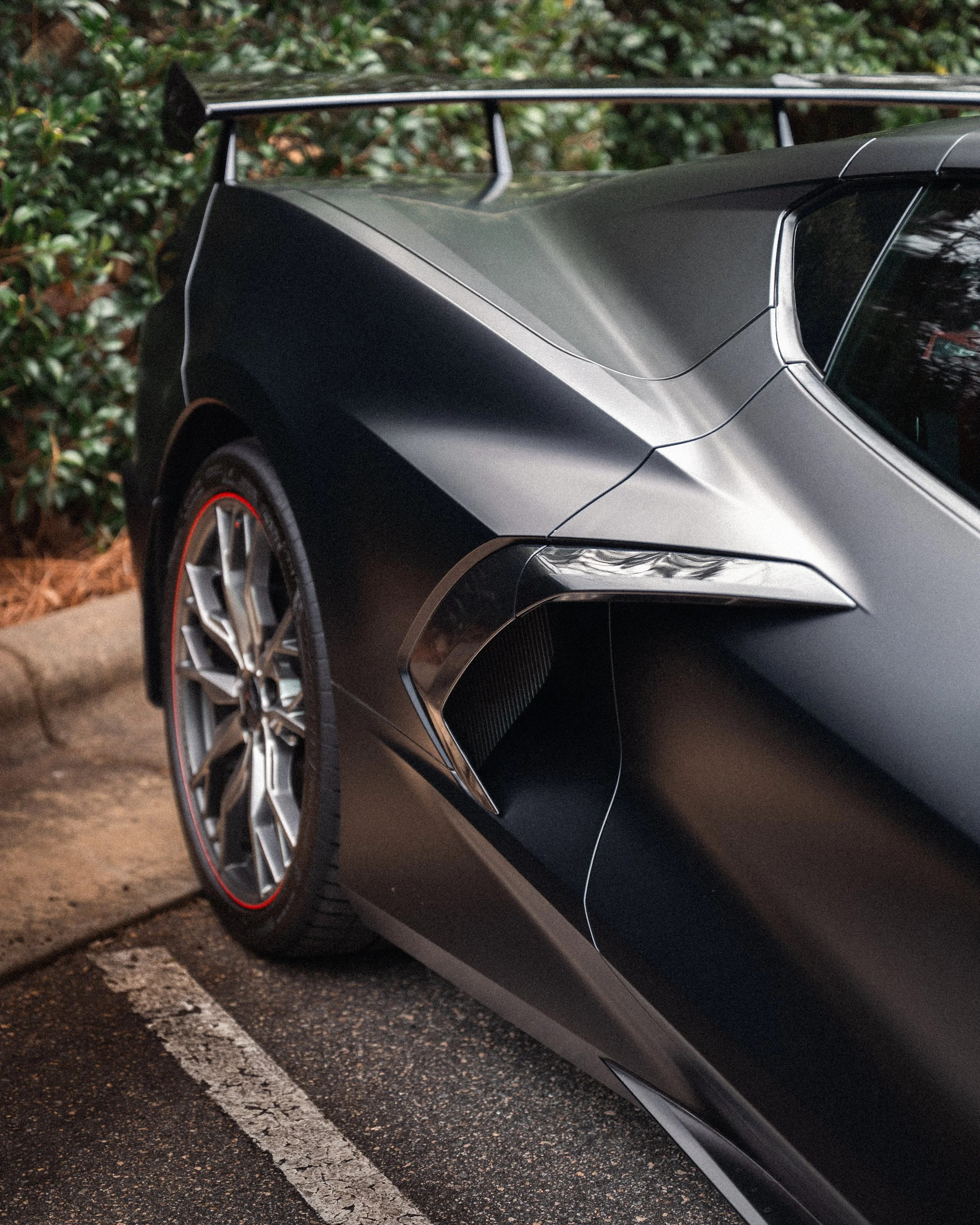 Close-up of the rear side of a sleek black sports car with a prominent rear wing, parked on a road with a cracked white parking line, greenery in the background.
