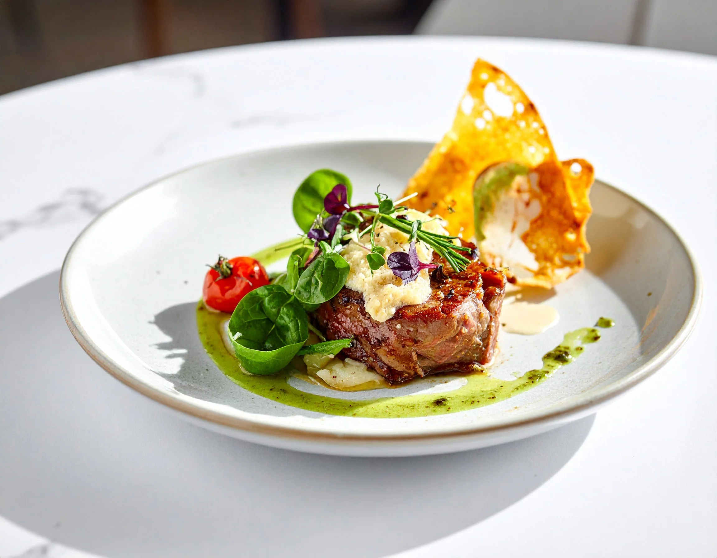 A white bowl containing a gourmet dish with a piece of cooked meat topped with cheese, a cherry tomato, microgreens, and a crispy cheese chip, with green sauce drizzled around the food.