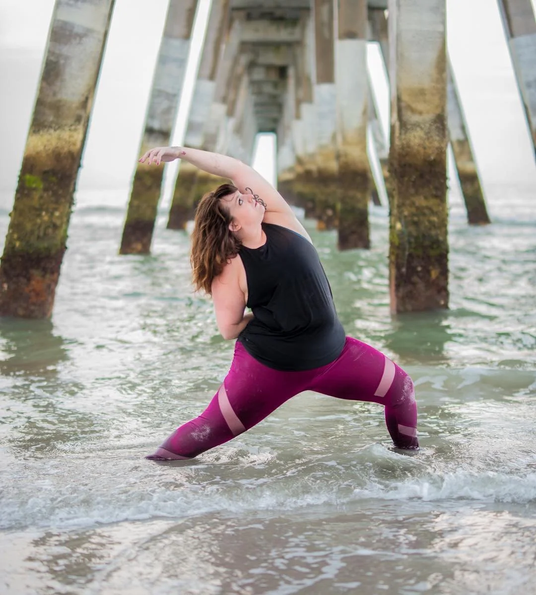A woman practicing yoga in the water under a pier, stretching with one arm overhead.