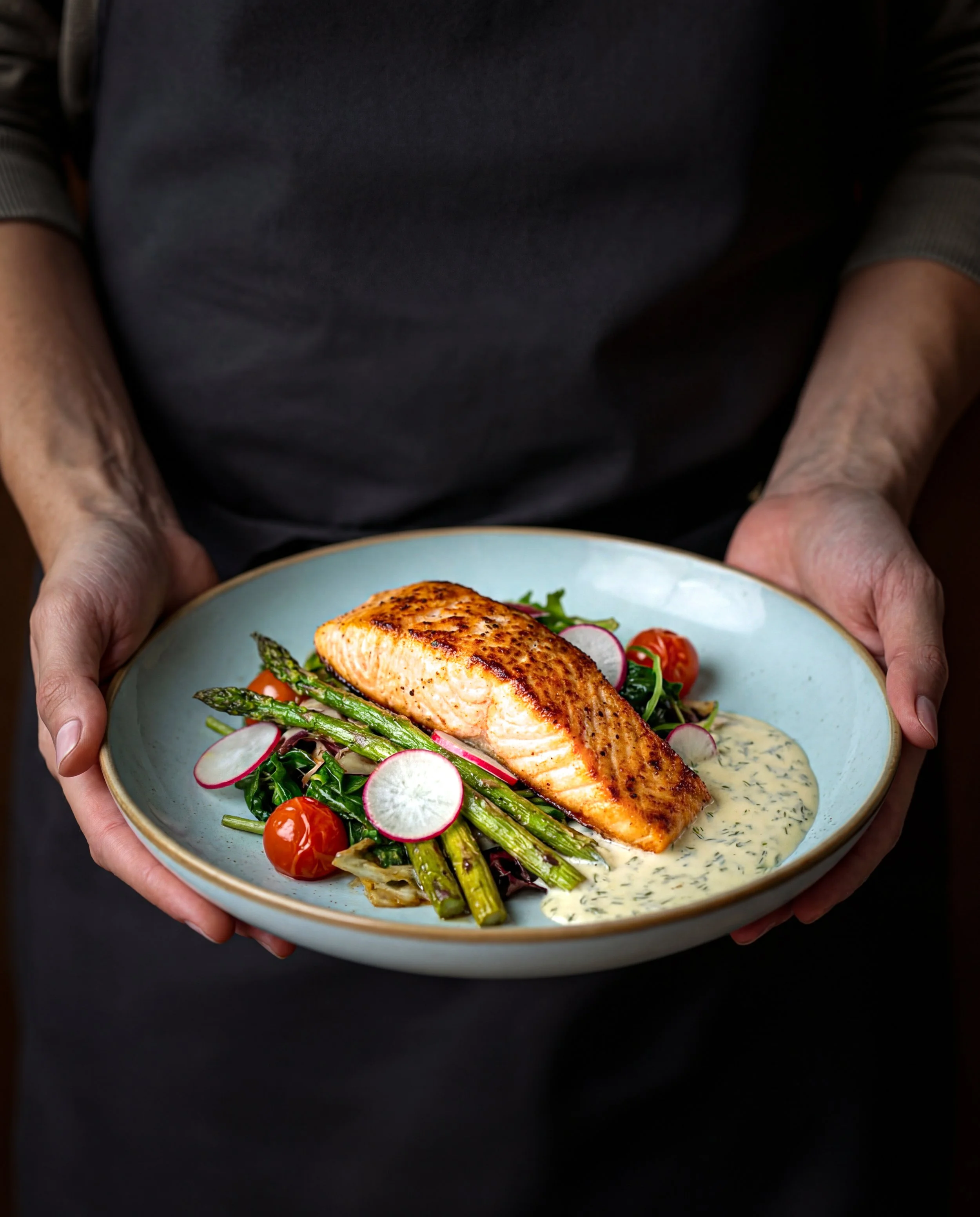 Person holding a plate with cooked salmon, asparagus, cherry tomatoes, radish slices, and a creamy herb sauce.