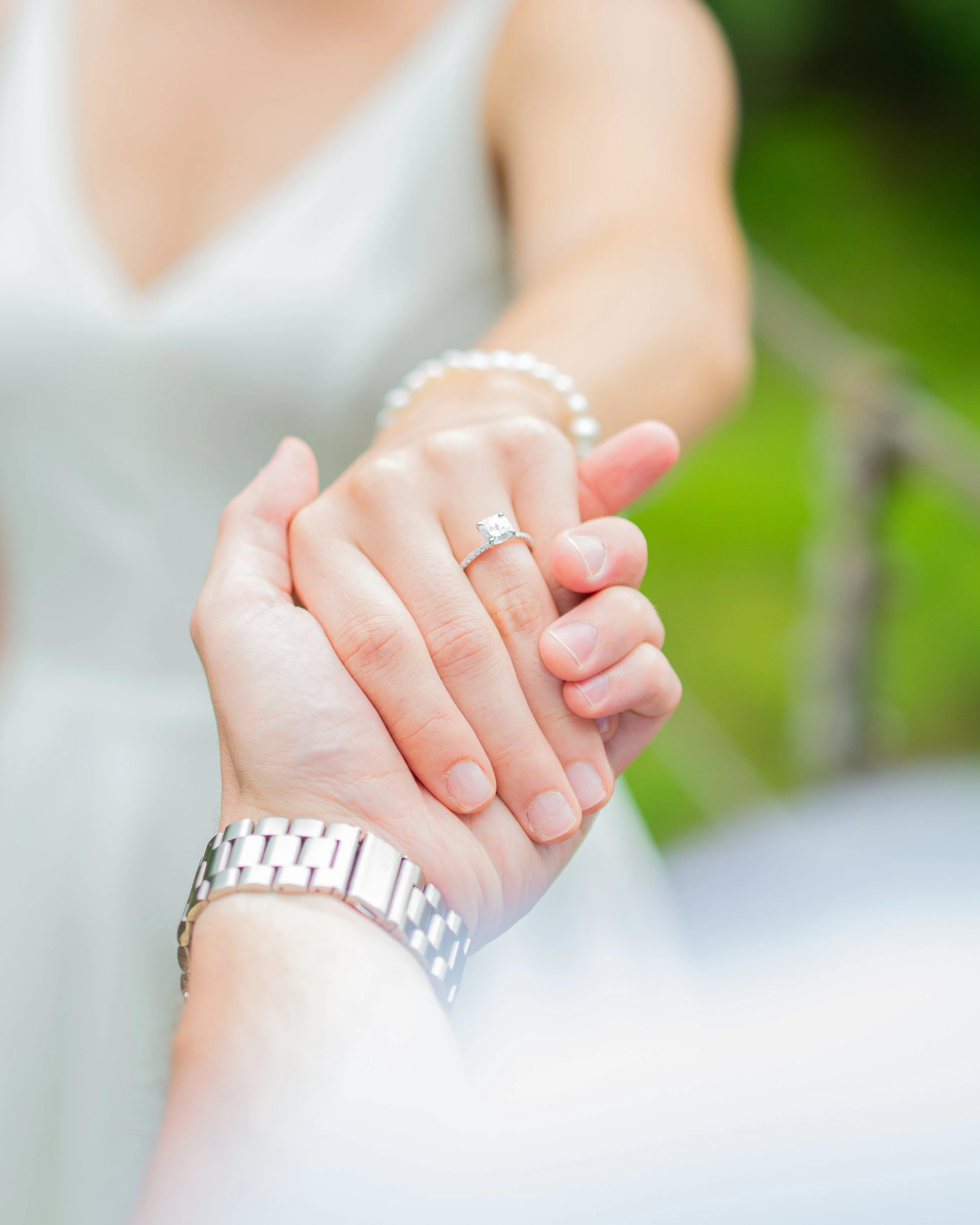 Close-up of a hand showing an engagement ring on the ring finger, with a woman in the background possibly showing her wedding band and bracelet.