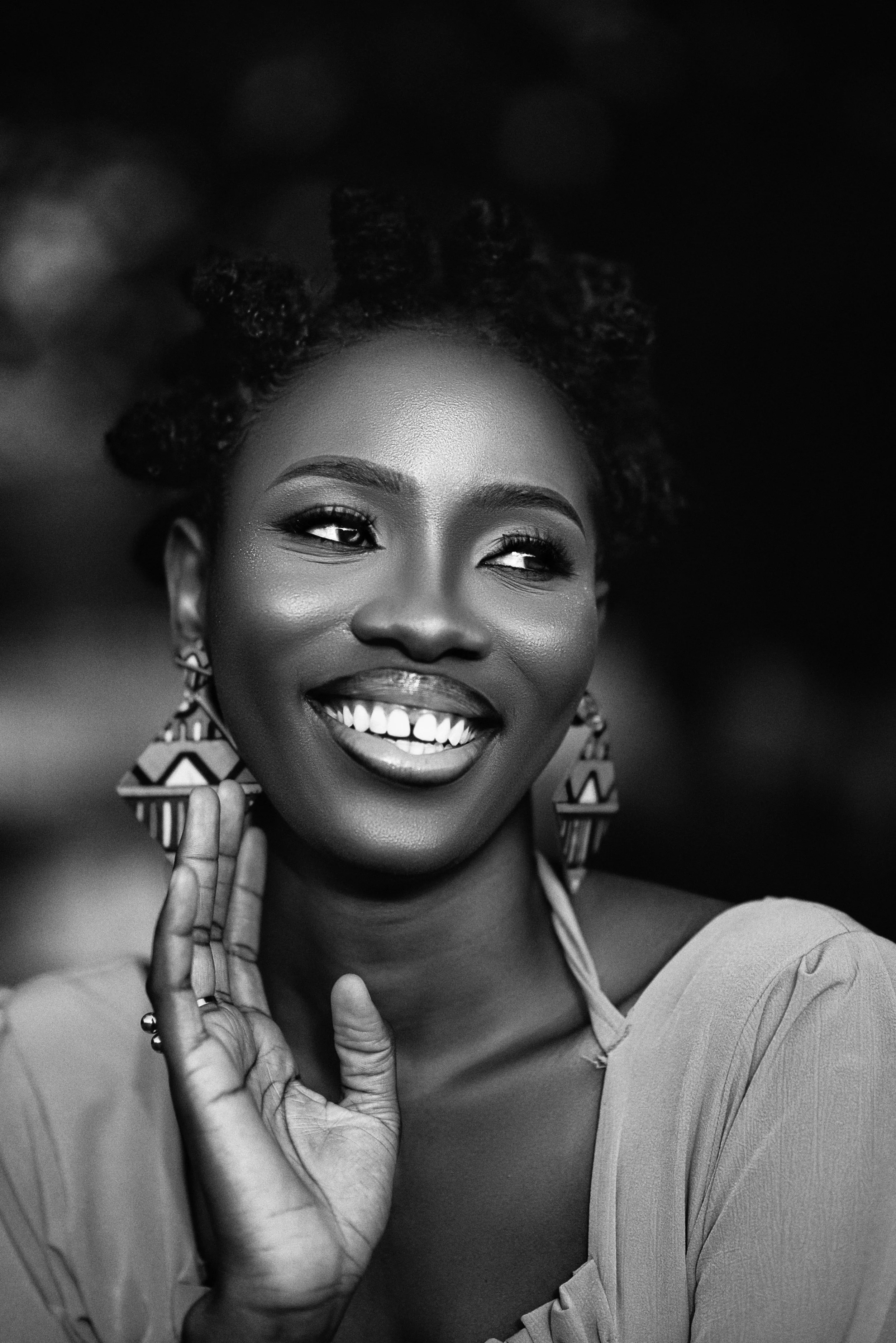 Black and white portrait of a smiling woman with curly hair, wearing earrings and a blouse, holding her hand near her face.