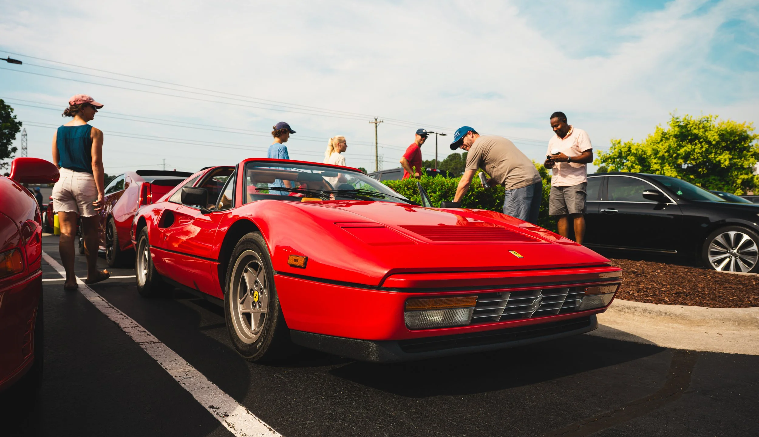 A group of people gathered around a bright red Ferrari sports car parked in a lot, with other vehicles and trees in the background on a sunny day.