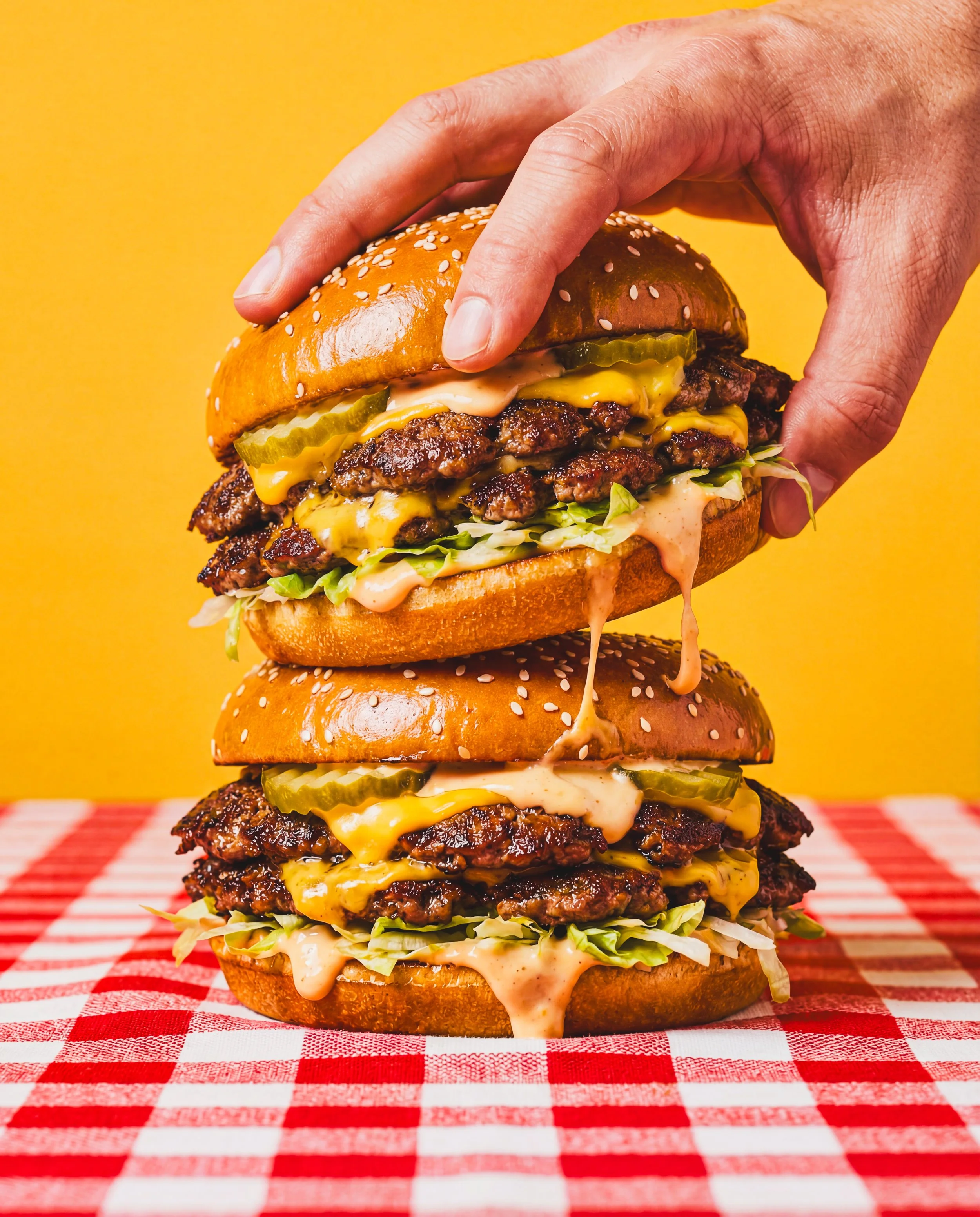 A hand stacking a double cheeseburger with lettuce, pickles, onions, and sauce on a red and white checkered tablecloth with a yellow background.