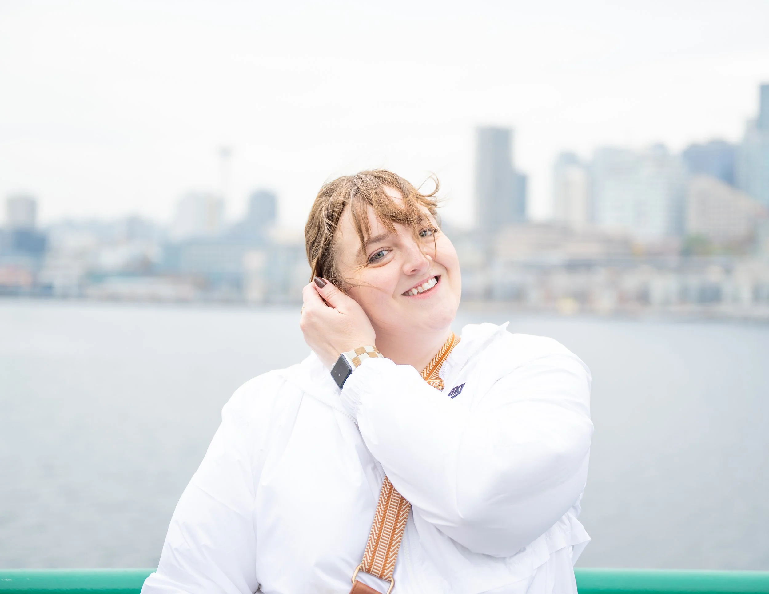 Woman with short brown hair smiling, wearing a white jacket and a smartwatch, outdoors near water with a city skyline in the background.