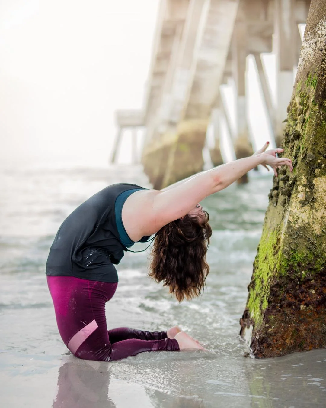 A woman practicing yoga on the beach, performing a pose with arms extended toward a moss-covered rock, near a wooden pier.