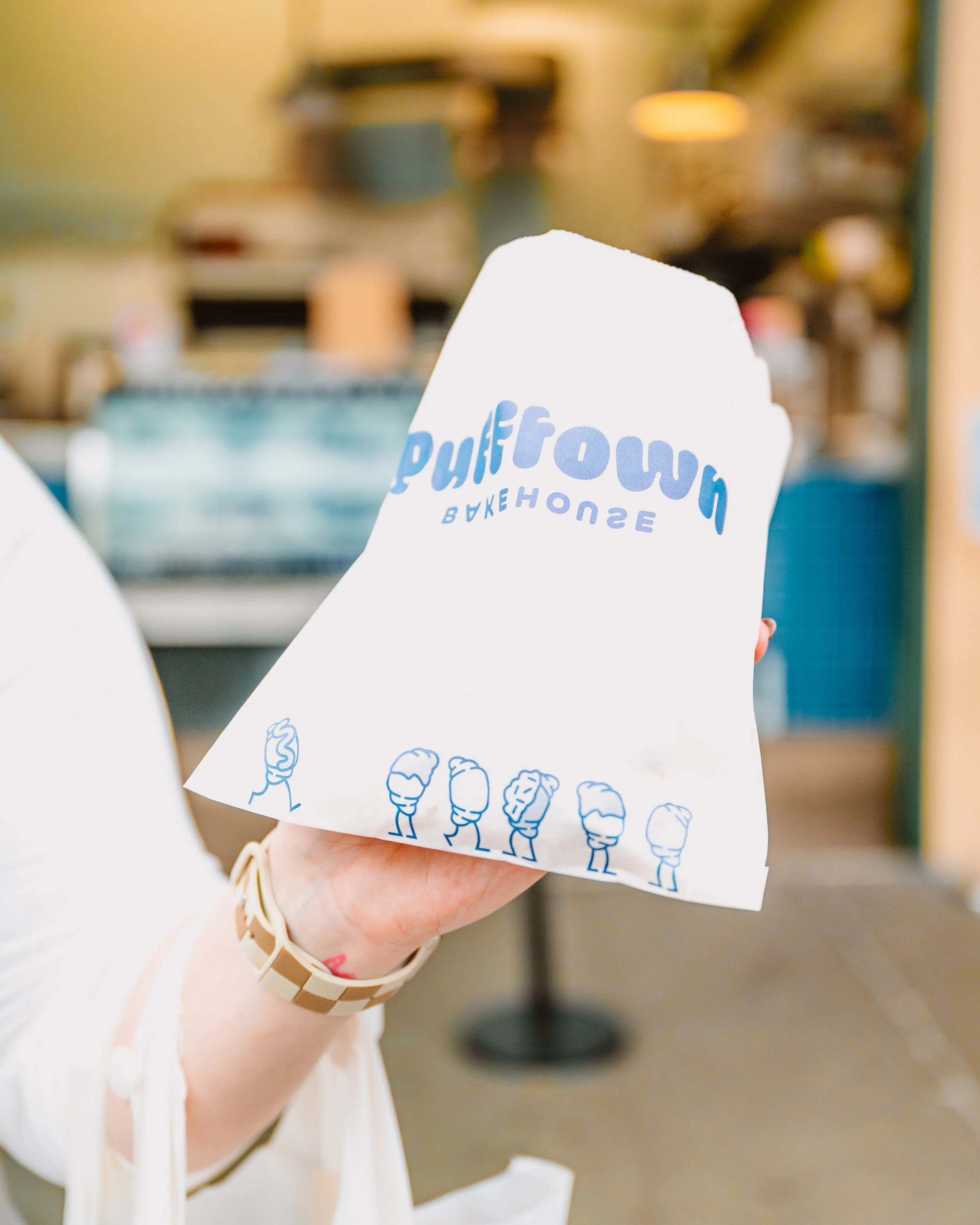 Person holding a white paper bag with blue text and illustrations inside a cafe.