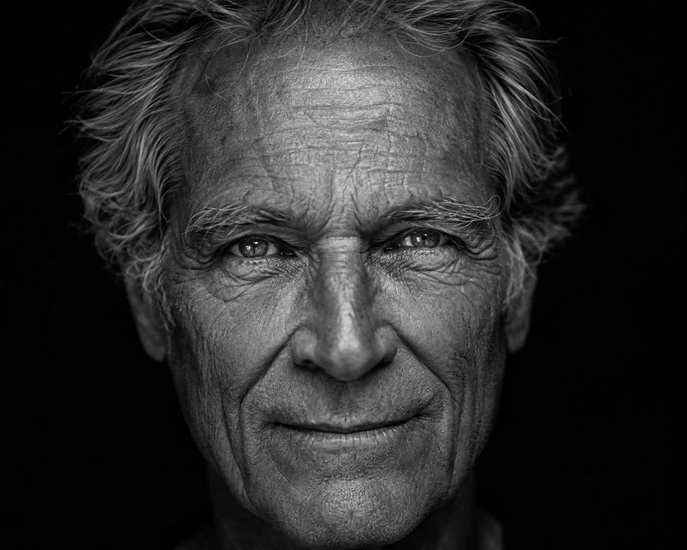 A close-up black and white portrait of an elderly man with wrinkles and gray hair, smiling softly.
