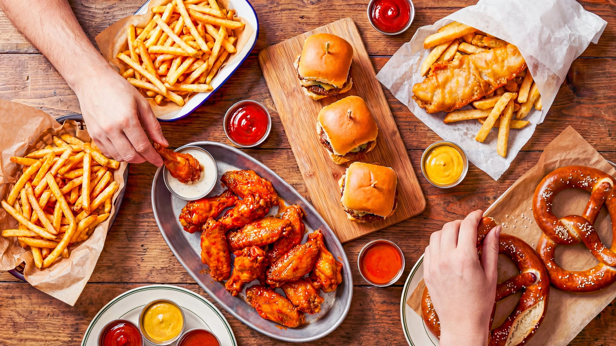 A variety of fast food on a wooden table including french fries, chicken wings, burgers, ketchup and mustard sauces, pretzels, and a person reaching for food.