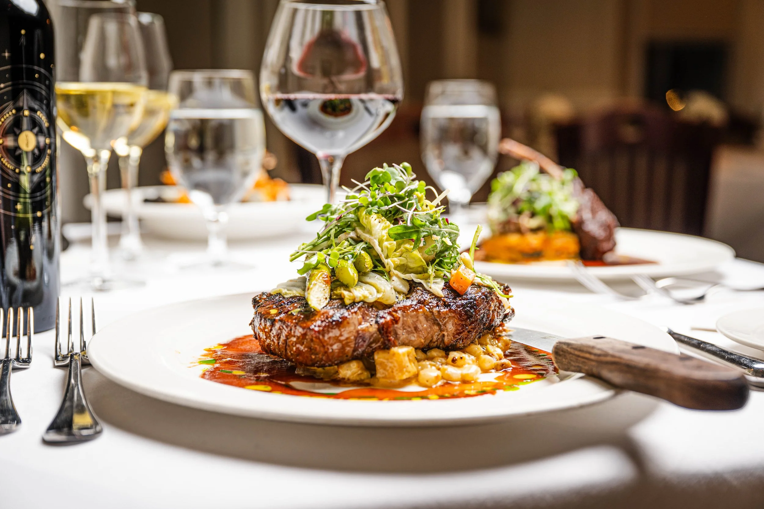 A plated dinner features a grilled steak topped with a vegetable medley and microgreens, served with sauce on a white plate. In the background, there are glasses of water and wine, a bottle of wine, and other dishes on a dining table.