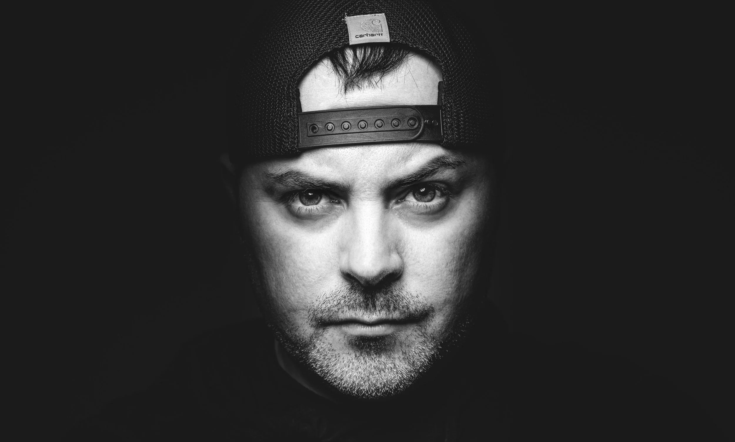 Black and white portrait of a man wearing a backward cap, staring directly at the camera.