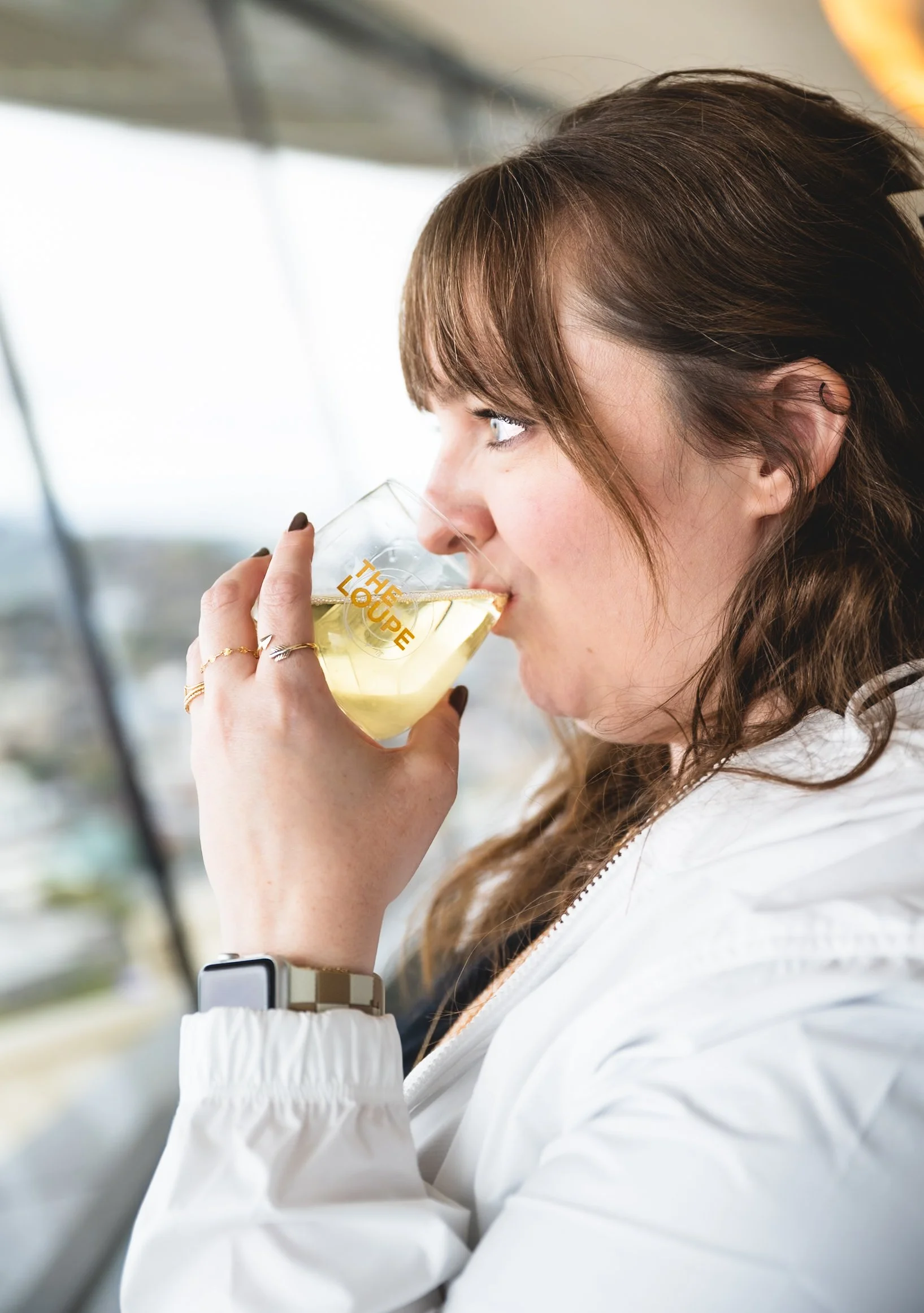 A woman with brown hair drinking white wine from a clear glass with yellow text that reads 'THE GROUP' in a bright indoor setting.