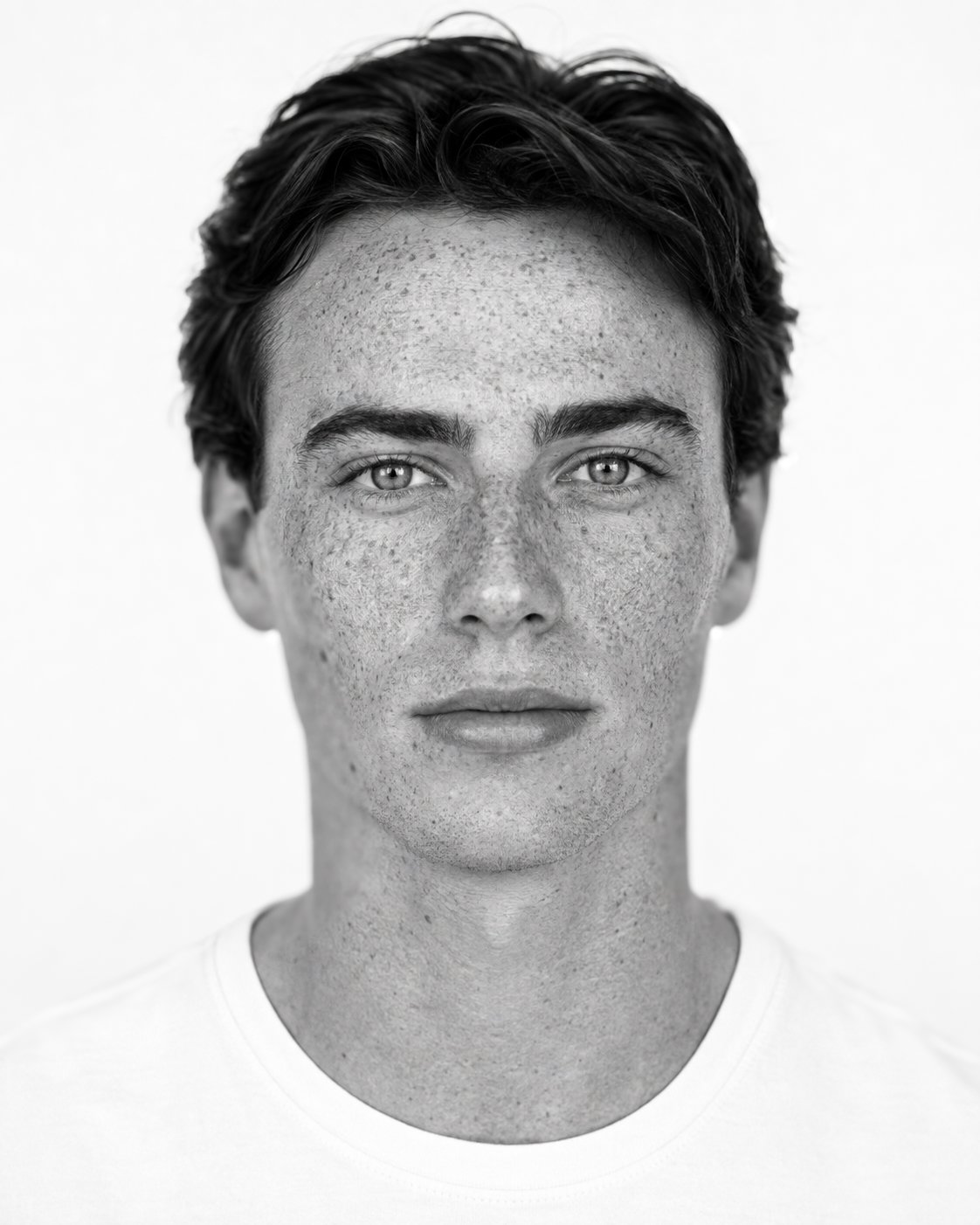 Black and white close-up portrait of a young person with short, dark hair, light skin with freckles, and light-colored eyes, looking directly at the camera.