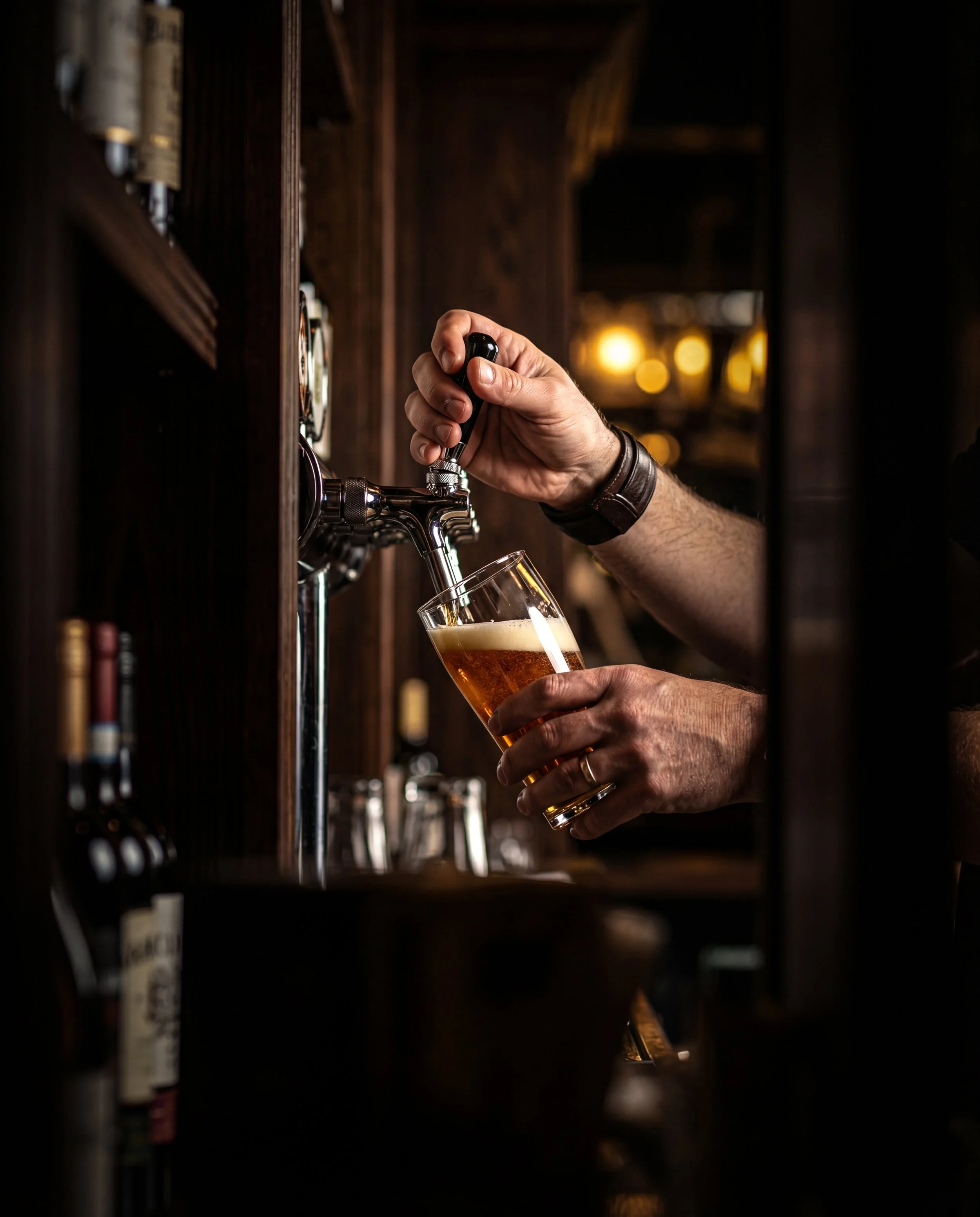 Person pouring beer from tap into glass at a bar with warm lighting.