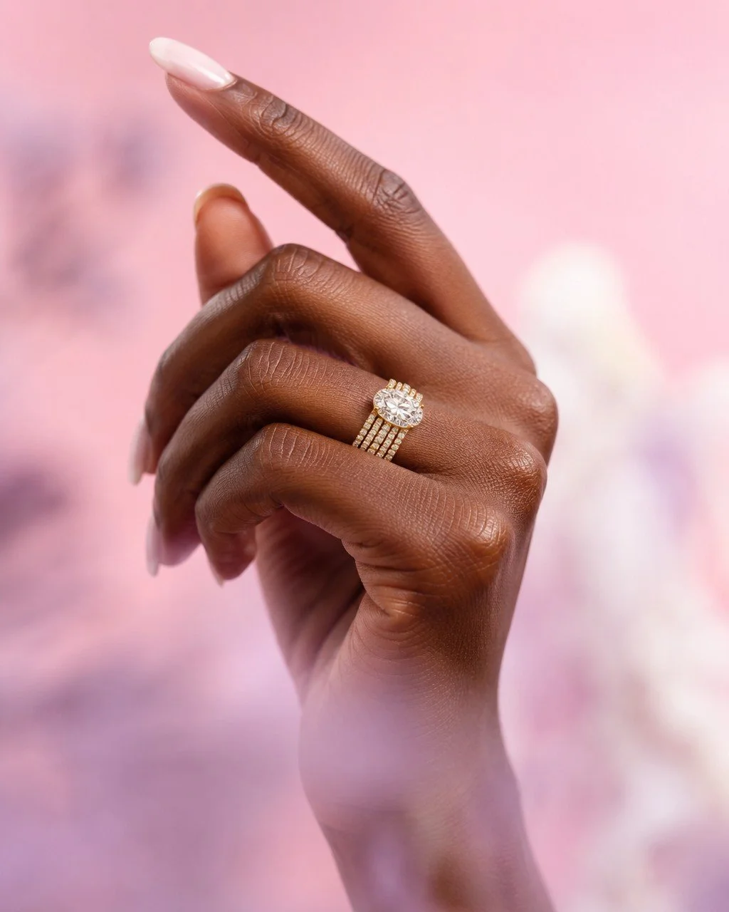 Close-up of a hand with dark skin, showing a diamond ring on the ring finger against a soft pink background.