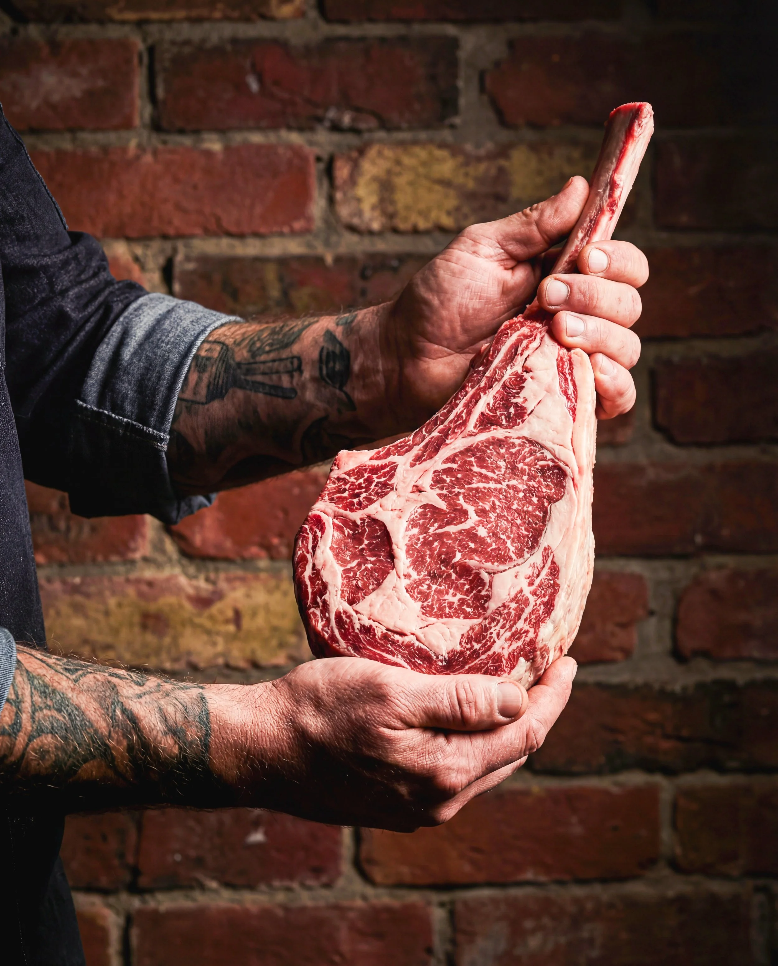 Person holding a large, marbled raw beef steak with a long bone against a brick wall background.
