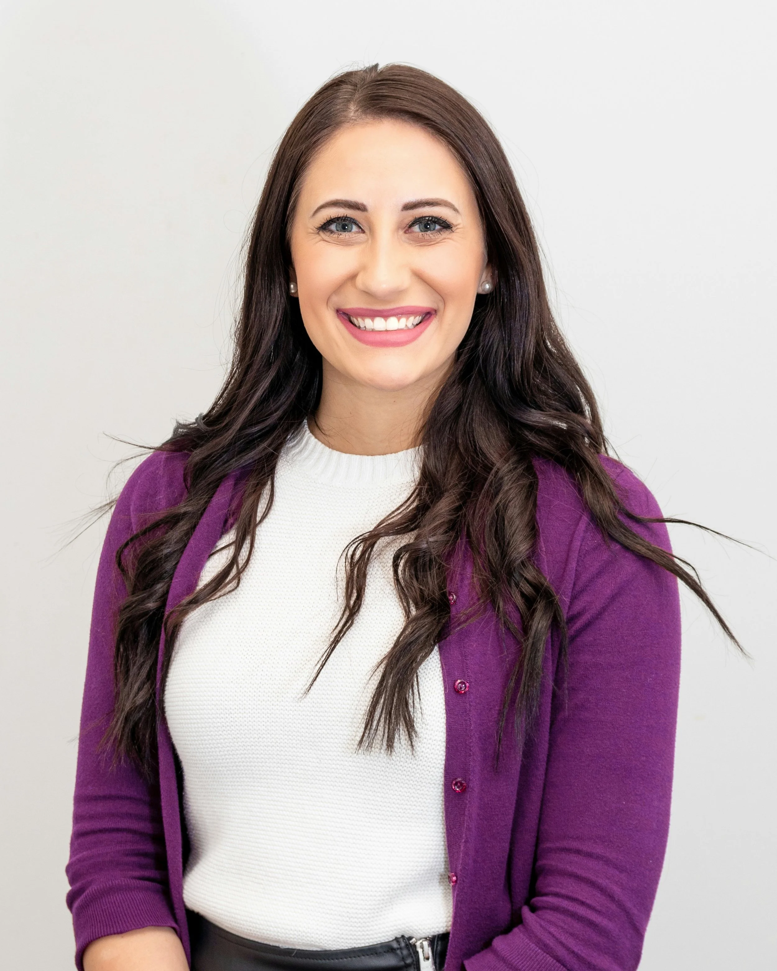 Portrait of a smiling woman with long dark hair, wearing a white top and a purple cardigan, against a plain white background.