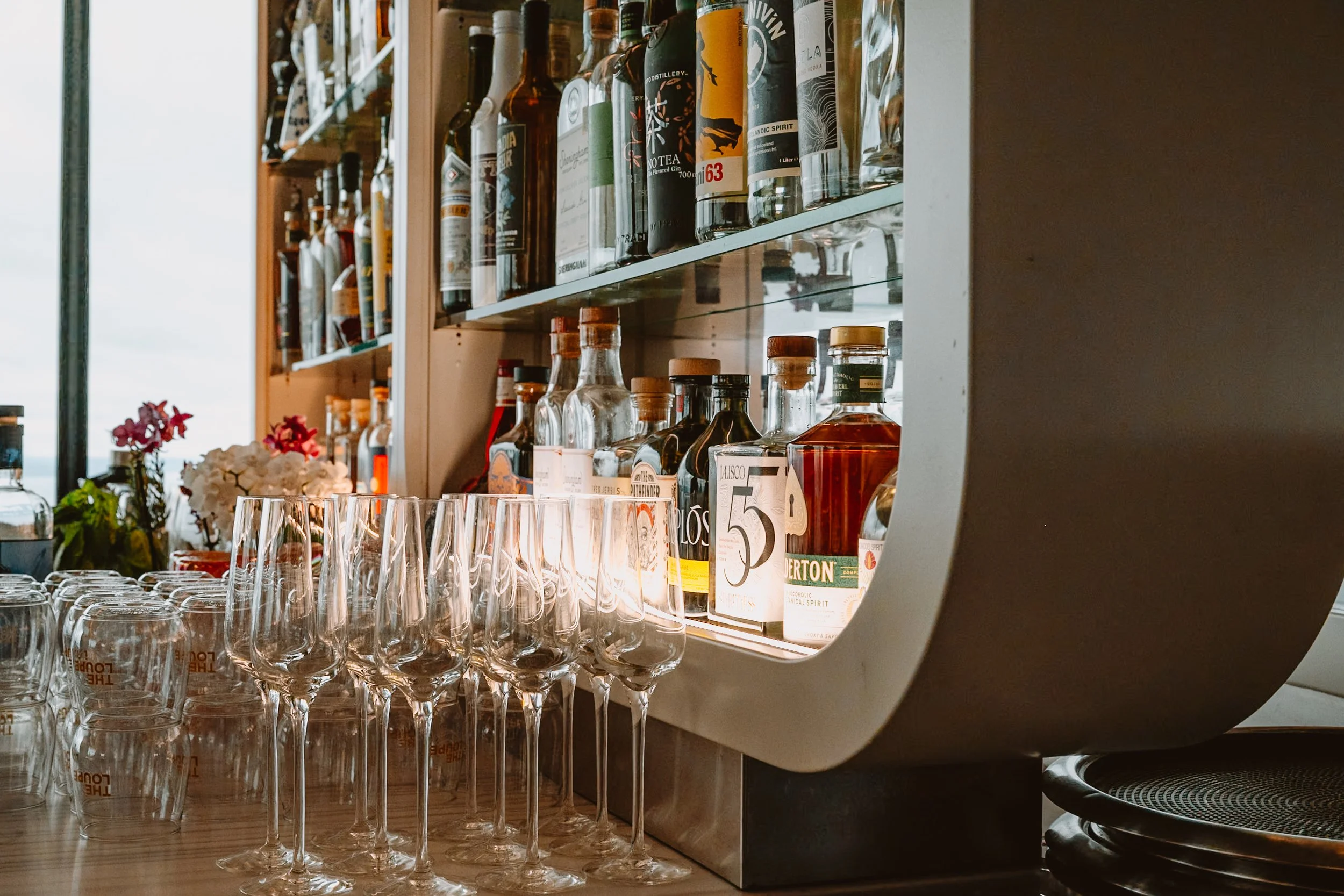A bar shelf holding various bottles of alcohol and liquor, with glasses arranged in front of the bottles. The shelf appears to be made of wood and glass, with a reflection visible underneath the bottles.