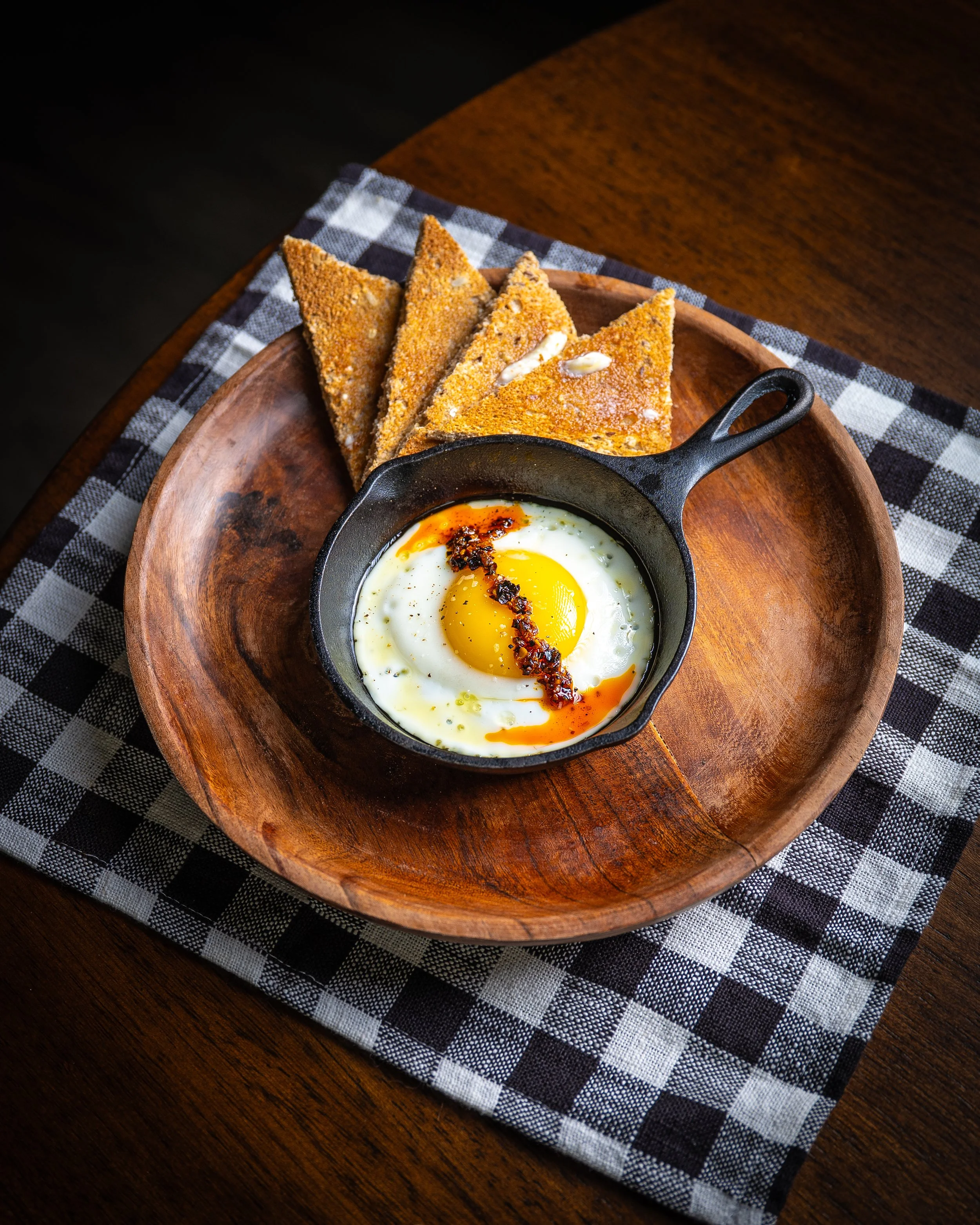 Fried egg in a skillet with red pepper flakes, served with toasted bread triangles on a wooden plate, all on a checkered black and white cloth.