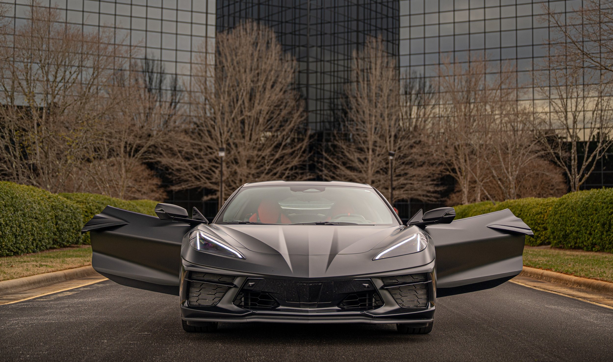 A sleek black sports car with open gull-wing doors is parked in front of a modern glass office building surrounded by leafless trees and greenery.