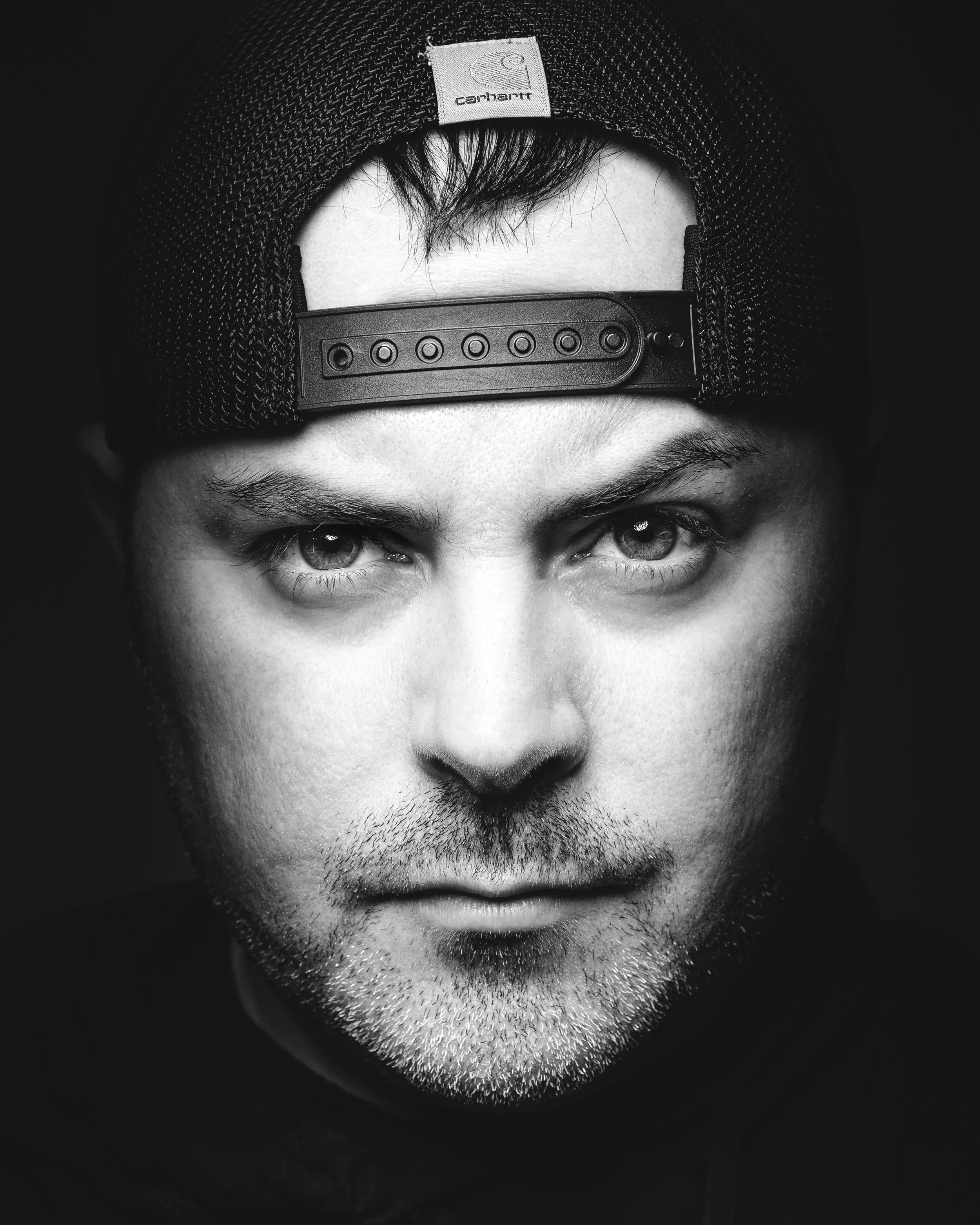 Close-up black and white photo of a man's face, wearing a backward baseball cap, with visible eyes, nose, and stubble.