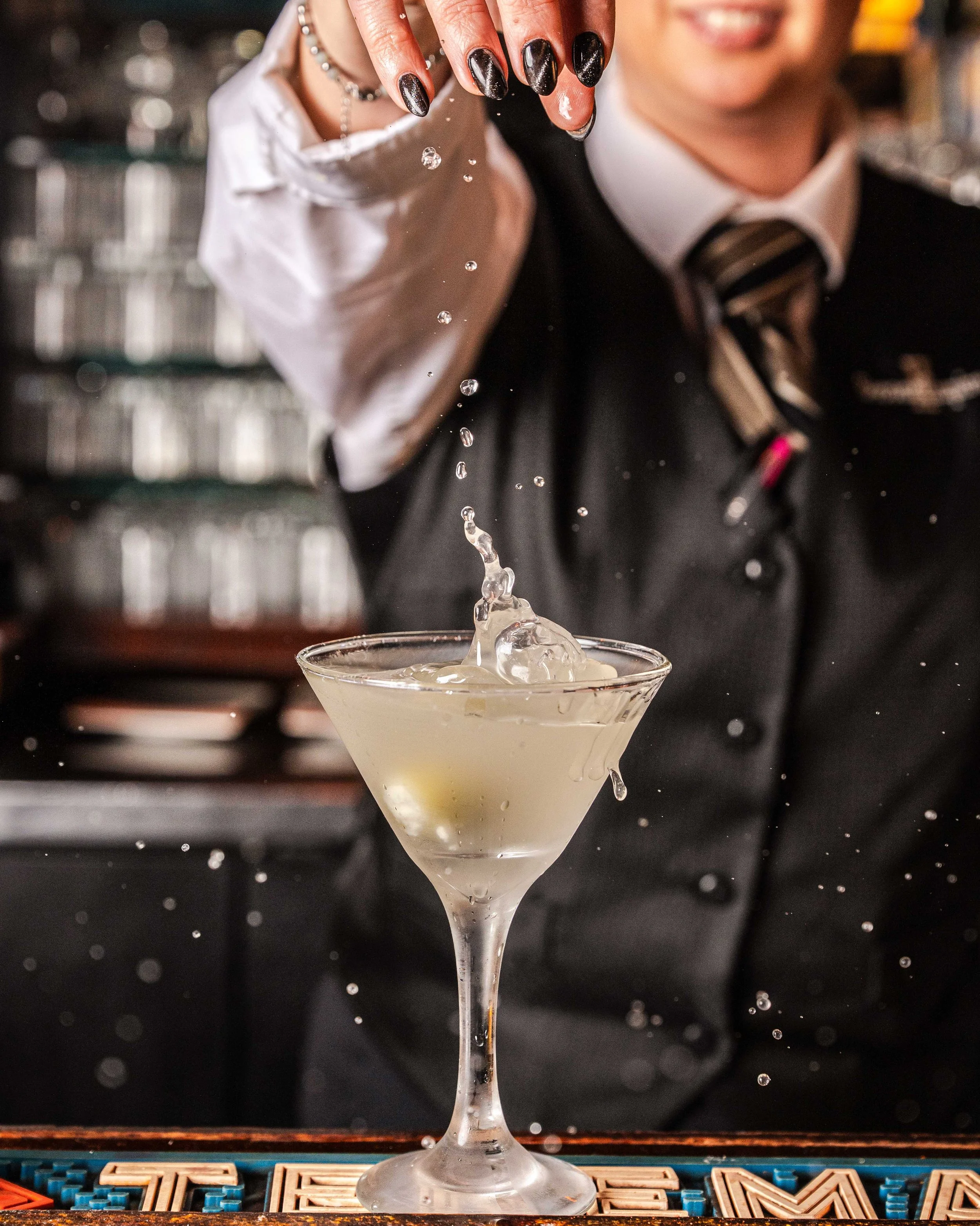 Bartender pouring a drink into a martini glass, with droplets splashing and a blurred background