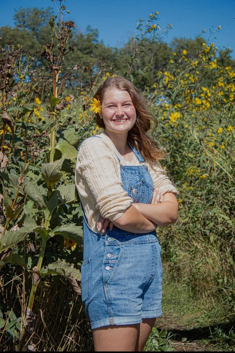 A young woman with curly hair and a sunflower behind her ear, standing in a field of sunflowers and yellow flowering bushes on a sunny day, smiling with her arms crossed.