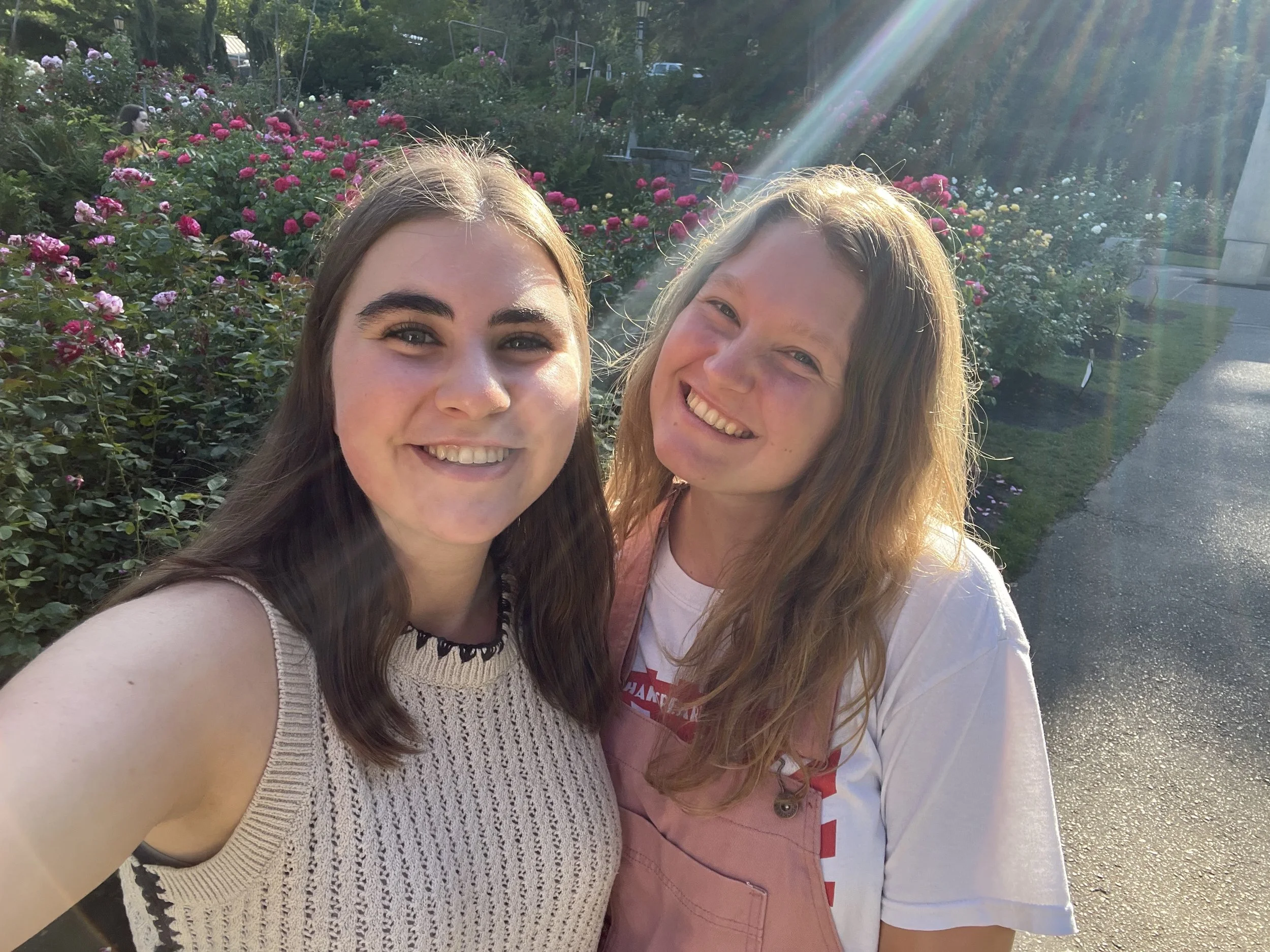 Two young women smiling outdoors with flowers and greenery in the background, sunlight and lens flare