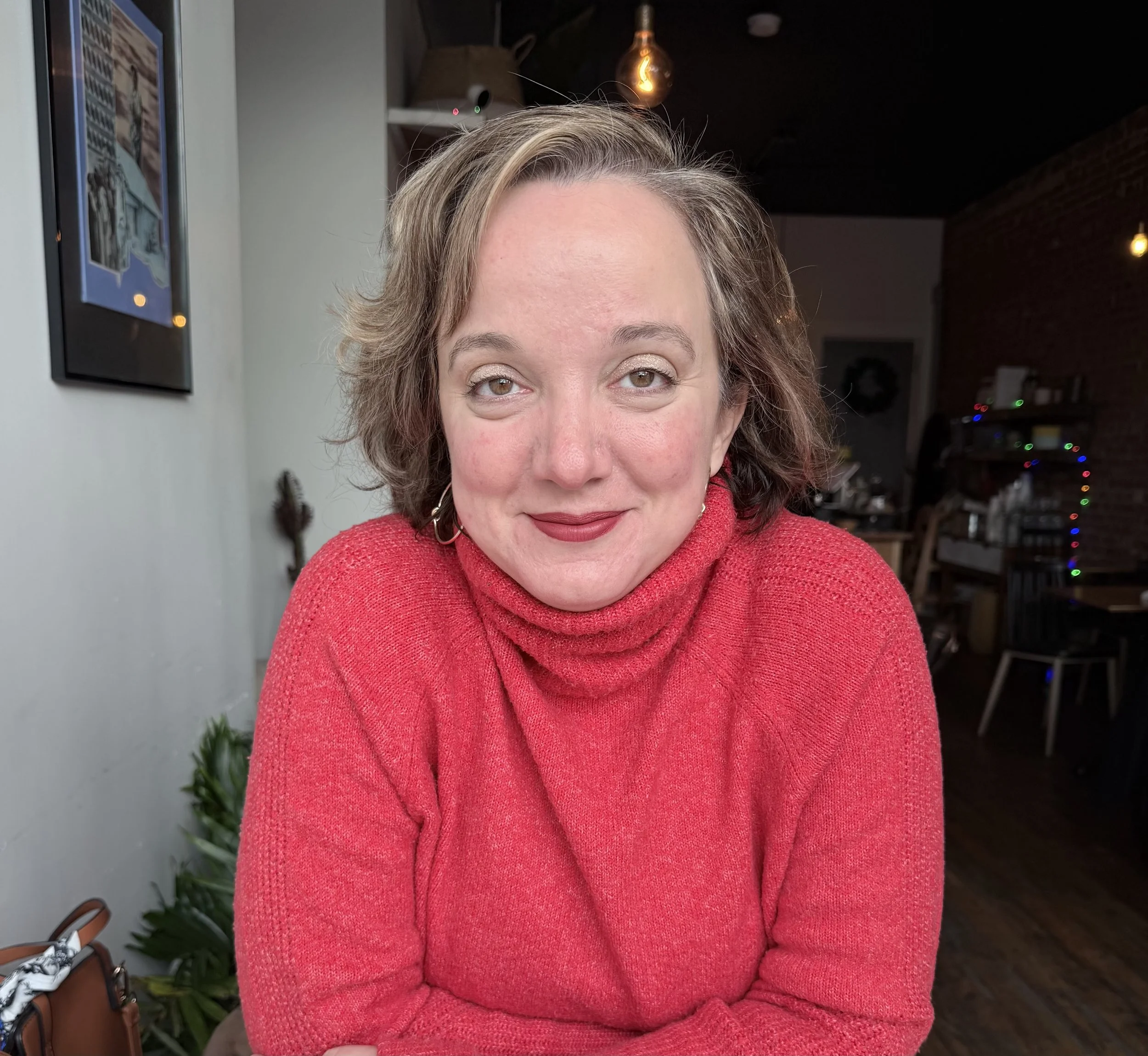 A woman with shoulder-length wavy hair wearing a red turtleneck sweater, sitting indoors with a brick wall and festive string lights in the background.
