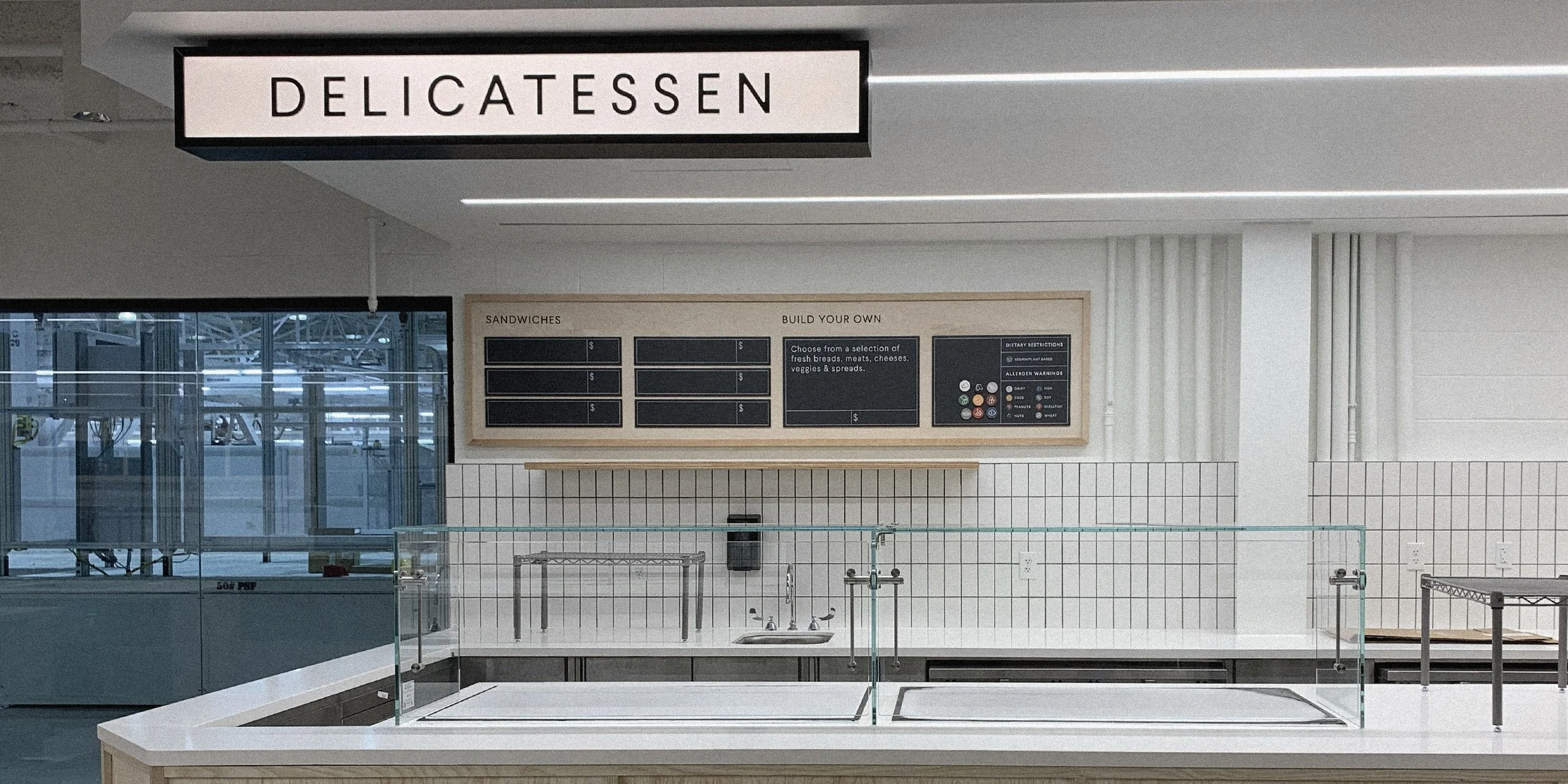 Empty delicatessen counter in a modern, minimalist grocery store with a black and white sign reading "DELICATESSEN" and a menu board behind the counter displaying sandwich options and build-your-own choices. The store has white tiled walls and bright